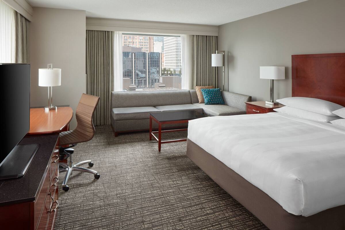 A hotel room with a bed, a desk with a TV, a sofa, a window with curtains, and a nightstand with a lamp.