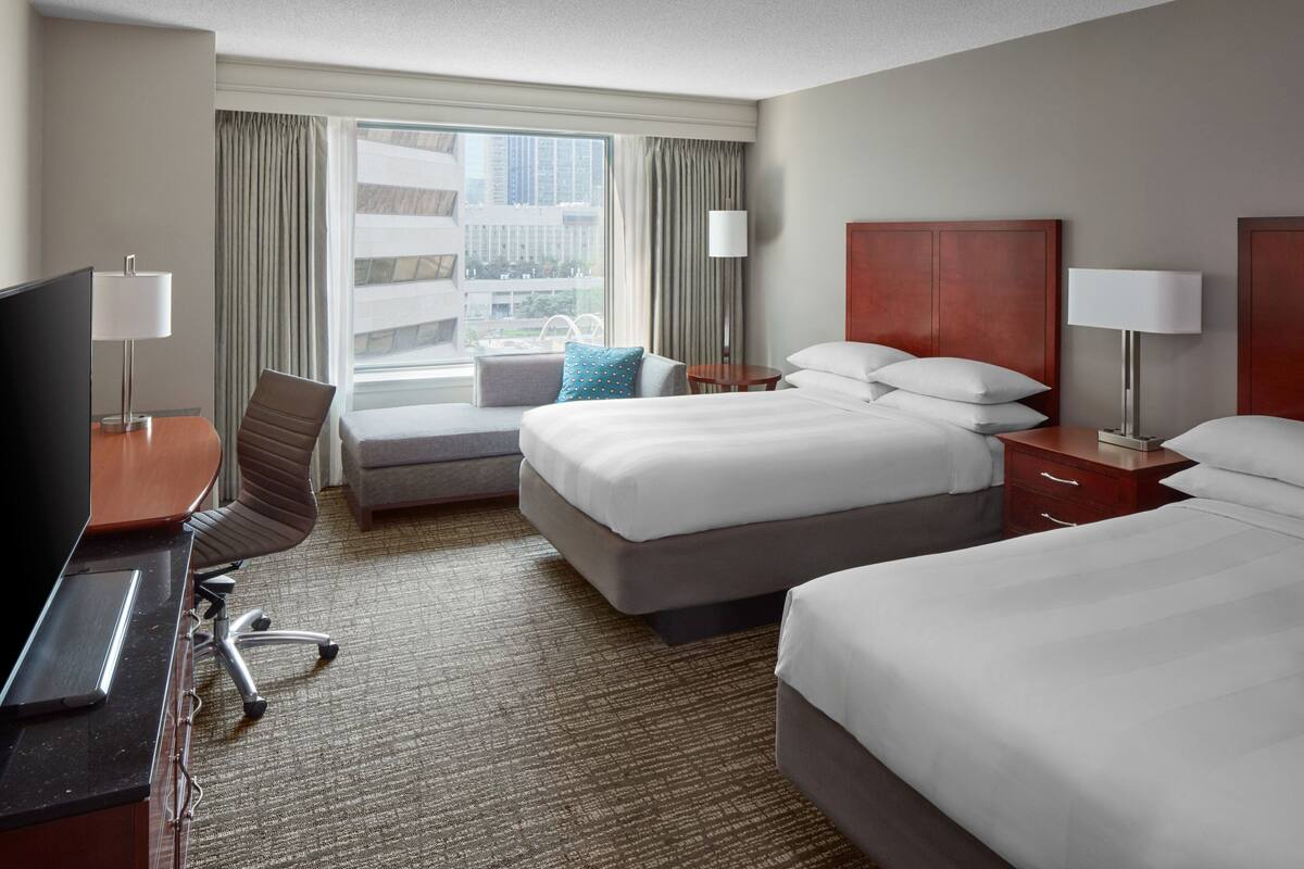 A hotel room with a bed, a desk with a TV, a chair, a small table, and a window with curtains.