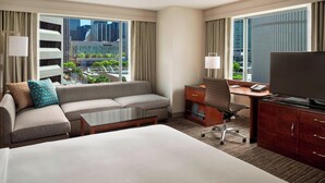 Habitación de hotel con una cama grande, un sofá, un escritorio, una silla y un televisor. Amplias ventanas con cortinas ofrecen vistas de la ciudad.