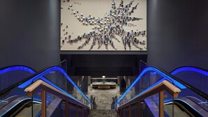 Un moderno vestíbulo de hotel con una gran instalación de arte abstracto en la pared, un mostrador de recepción central y una escalera con barandillas de vidrio.