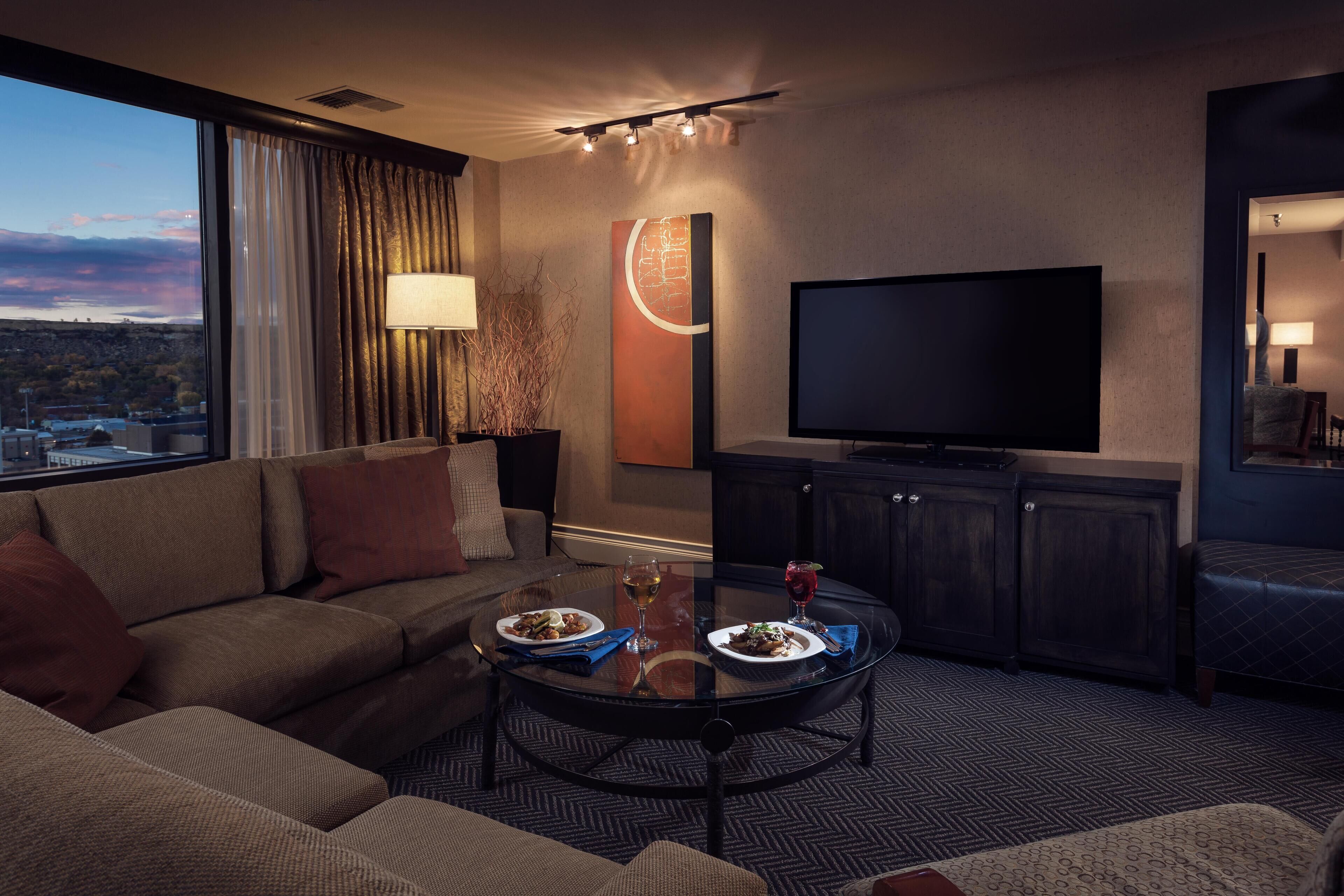 A modern hotel room with a large window, a flat-screen TV, a round glass coffee table, a sofa, and a cabinet.