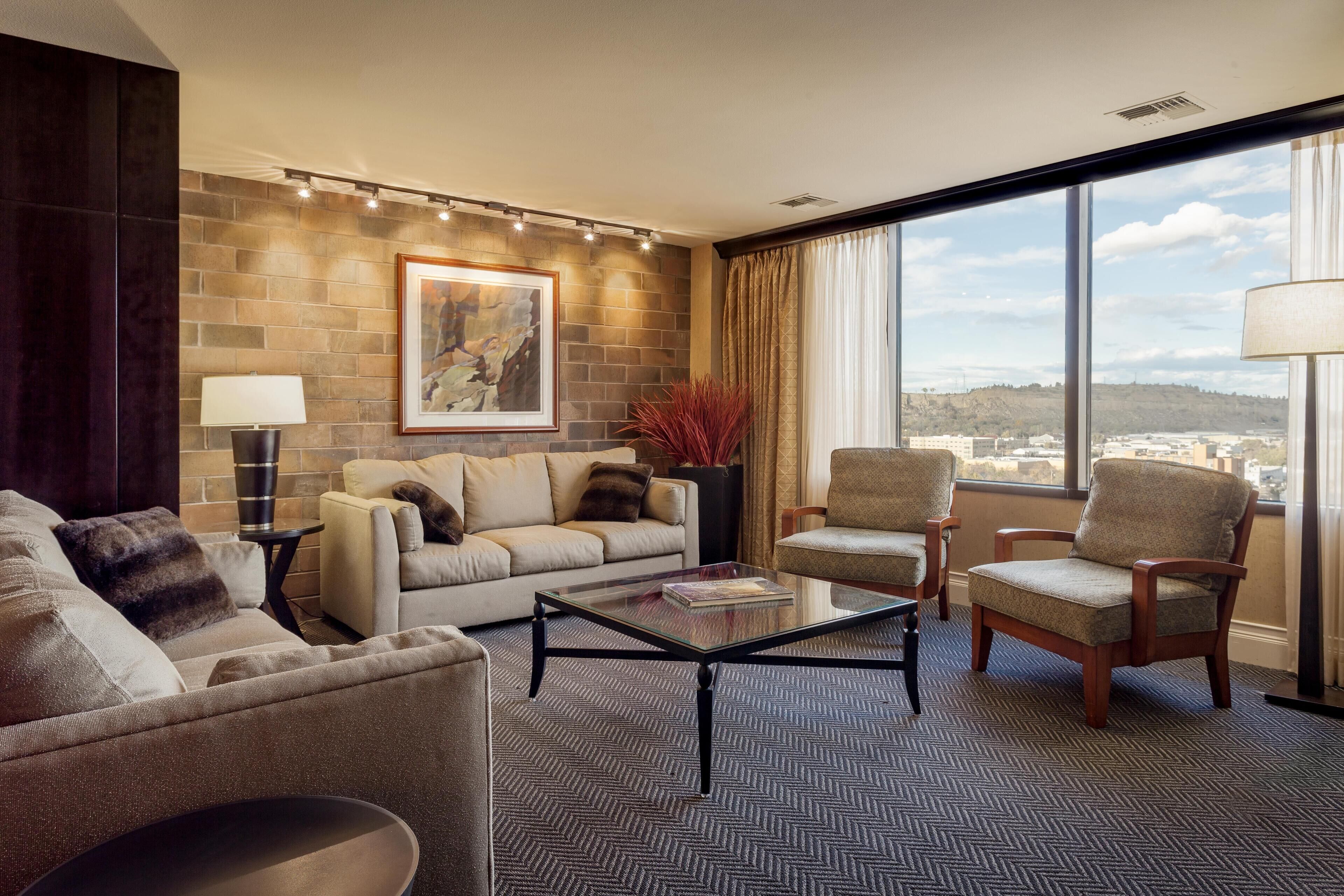 A modern living room with a sofa, armchairs, a coffee table, and a large window offering a city view.