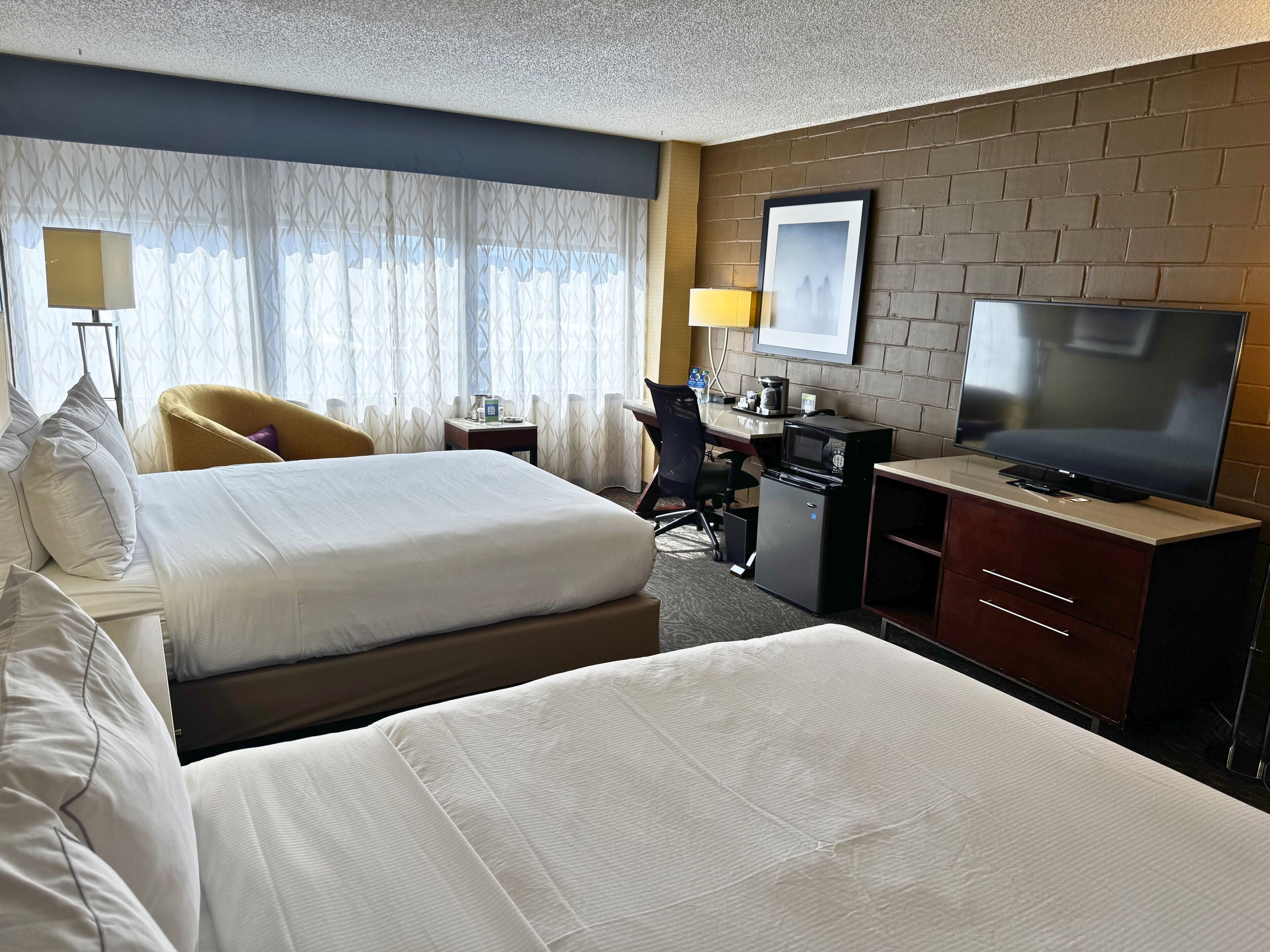 Habitación de hotel con una cama grande, un escritorio con computadora, televisor y ventana con cortinas.