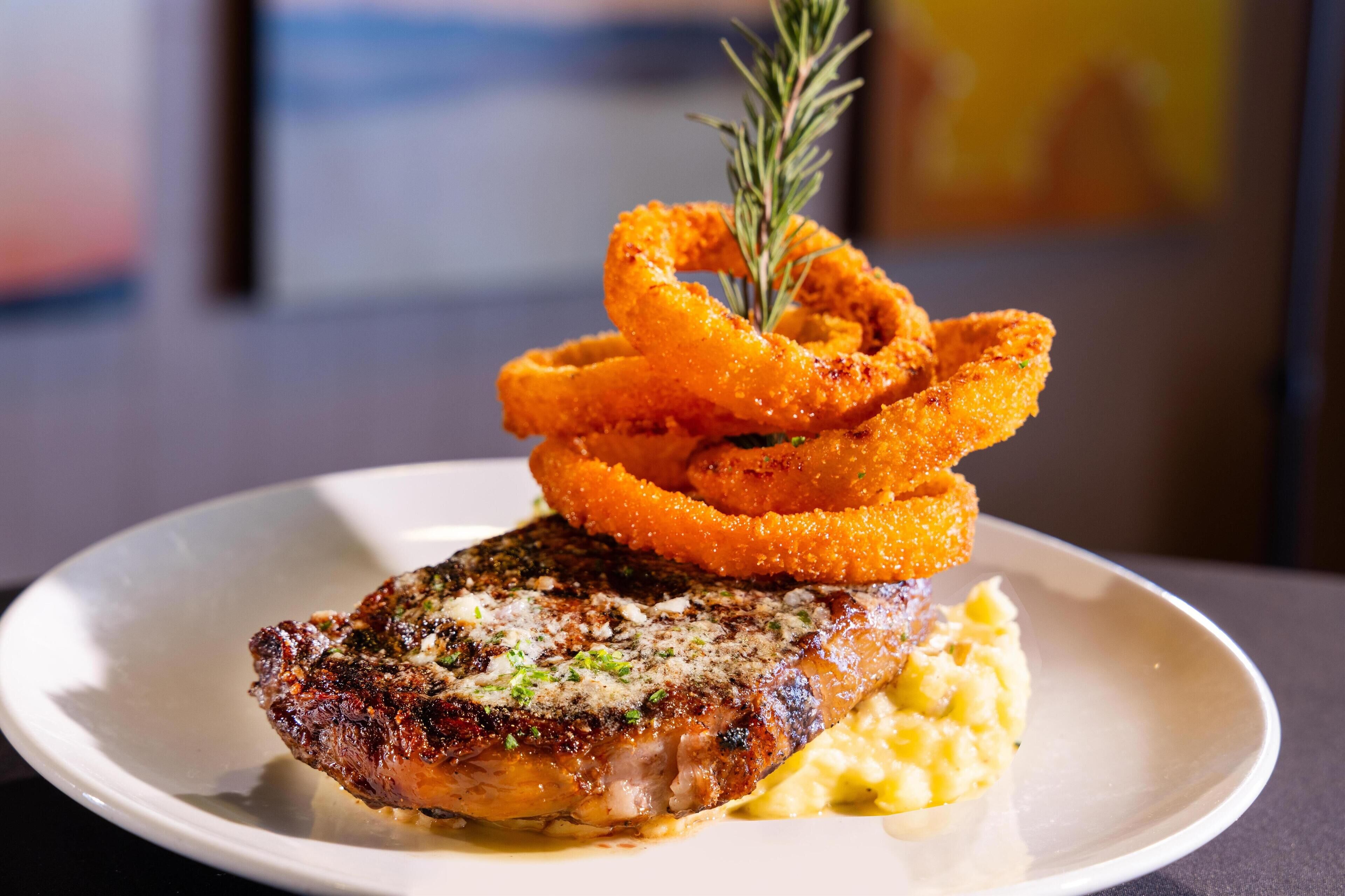 A plate with a grilled steak, onion rings, and mashed potatoes.
