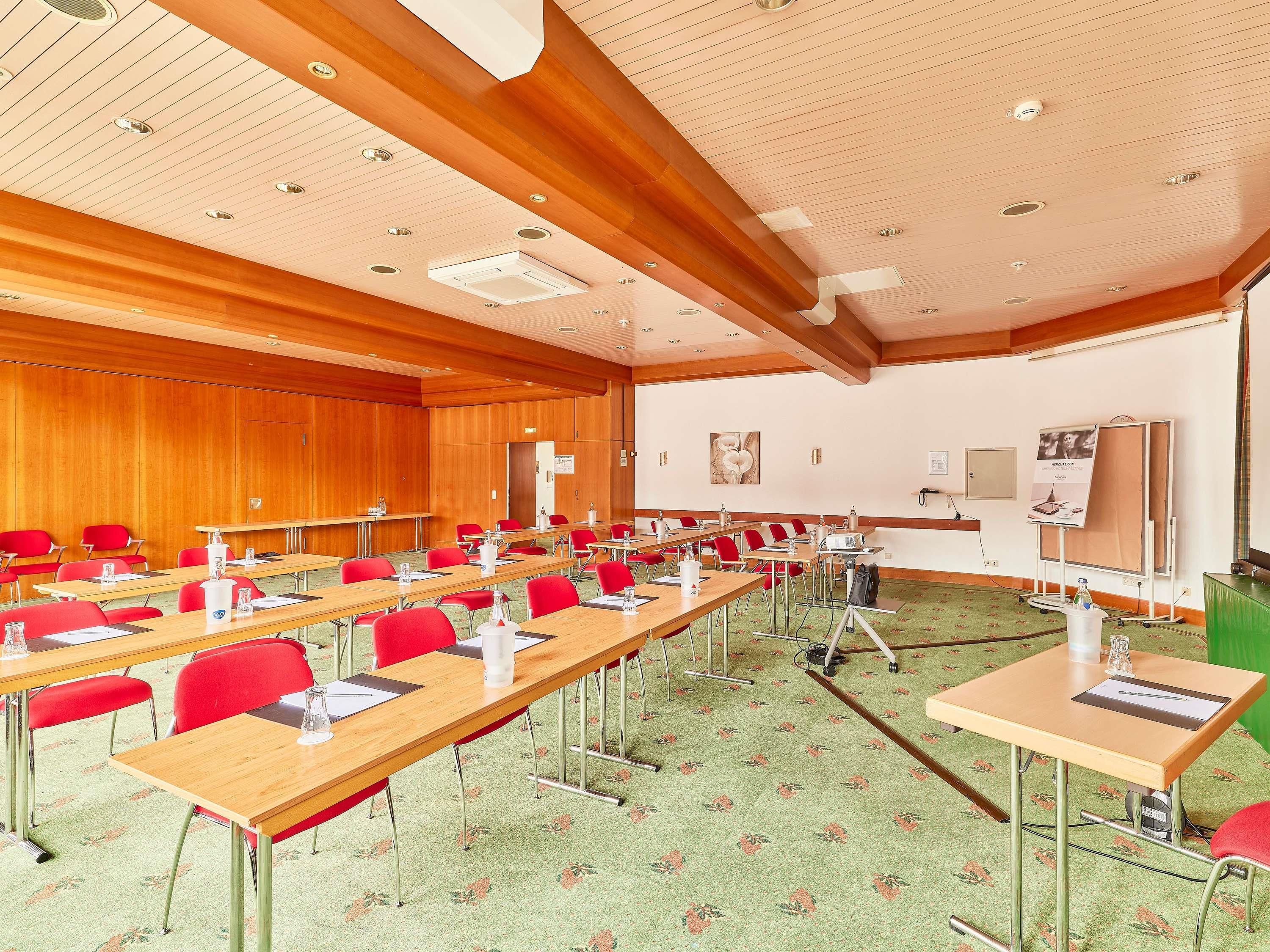 A conference room with long tables, red chairs, a green carpet, wooden panel walls, and ceiling lights.