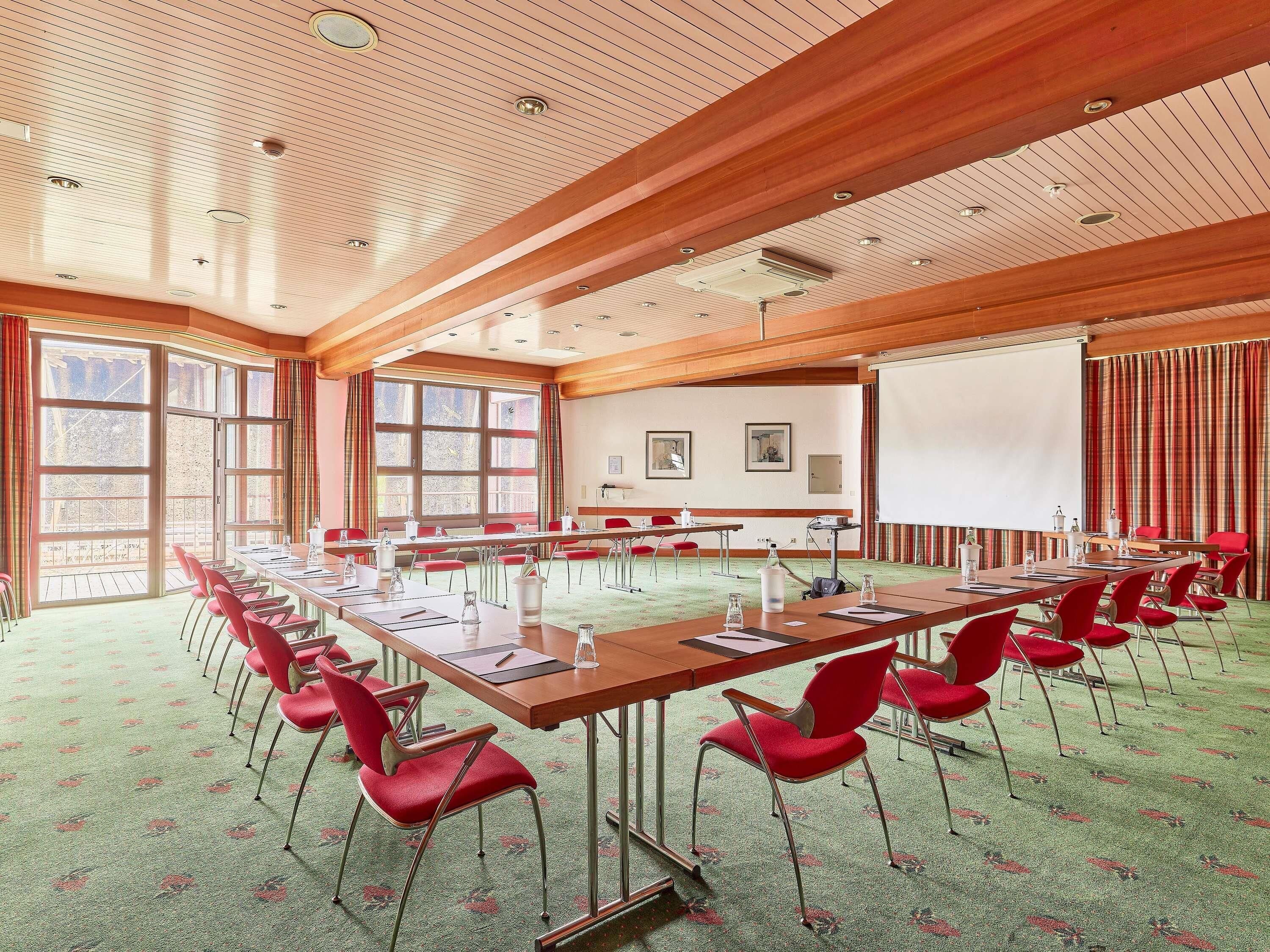 Un salón de conferencias con una larga mesa, sillas rojas, una pantalla de proyector y ventanas amplias.