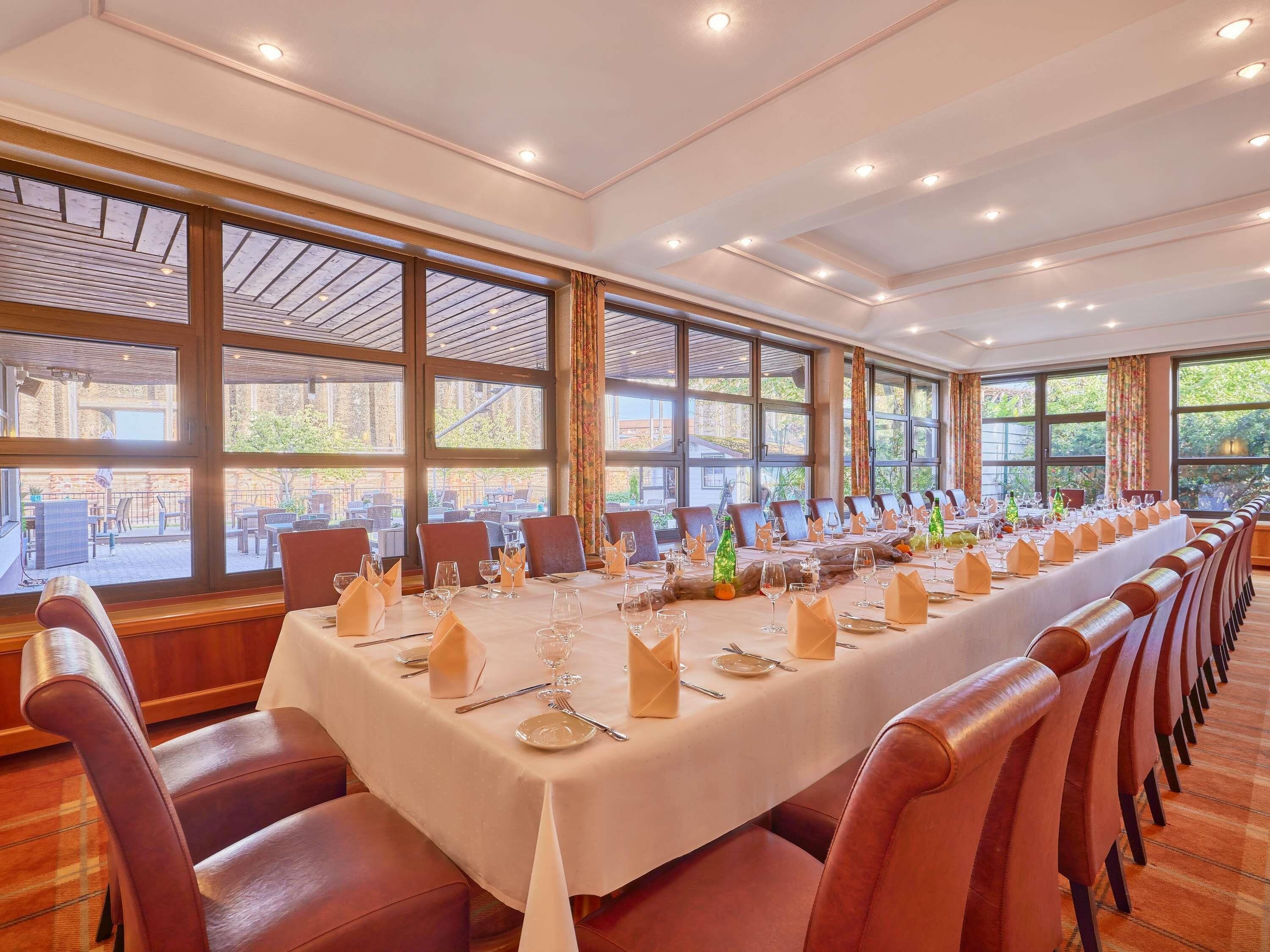 Una larga mesa de comedor preparada para una comida, con sillas, ventanas amplias y vistas al exterior.