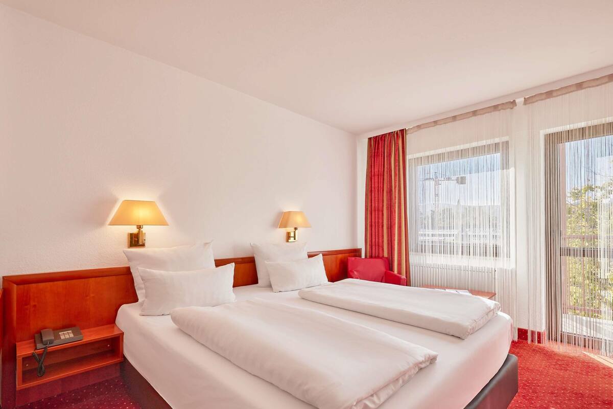 Hotelkamer met twee bedden, een houten hoofdbord, nachtkastjes met lampen, een rode stoel en een groot raam met gordijnen.