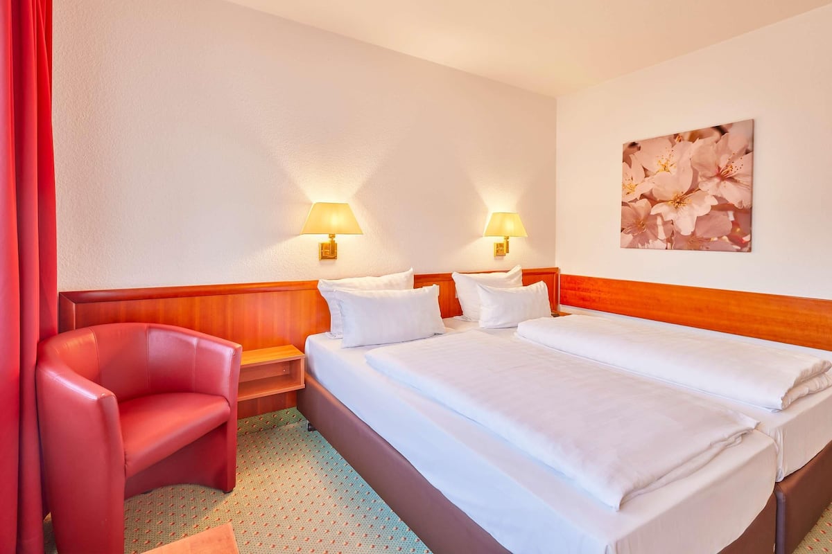 A hotel room with a double bed, two bedside lamps, a red armchair, and a wall-mounted picture of flowers.