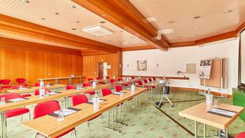 A conference room with long tables, red chairs, a green carpet, wooden panel walls, and ceiling lights.