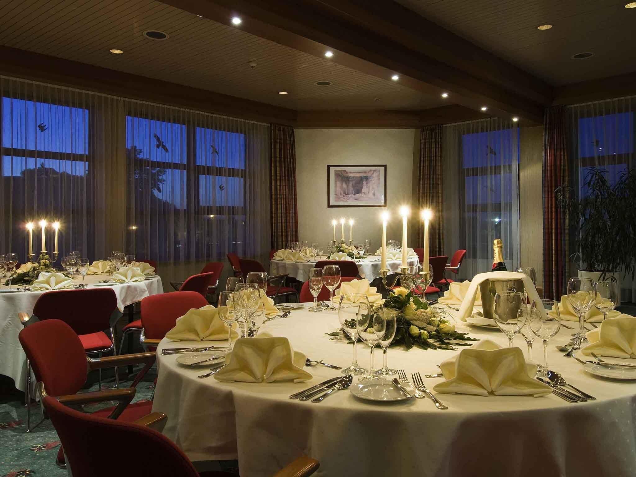 Une salle à manger élégante, avec des tables rondes dressées pour un repas, agrémentée de nappes blanches, de bougies et de chaises rouges.