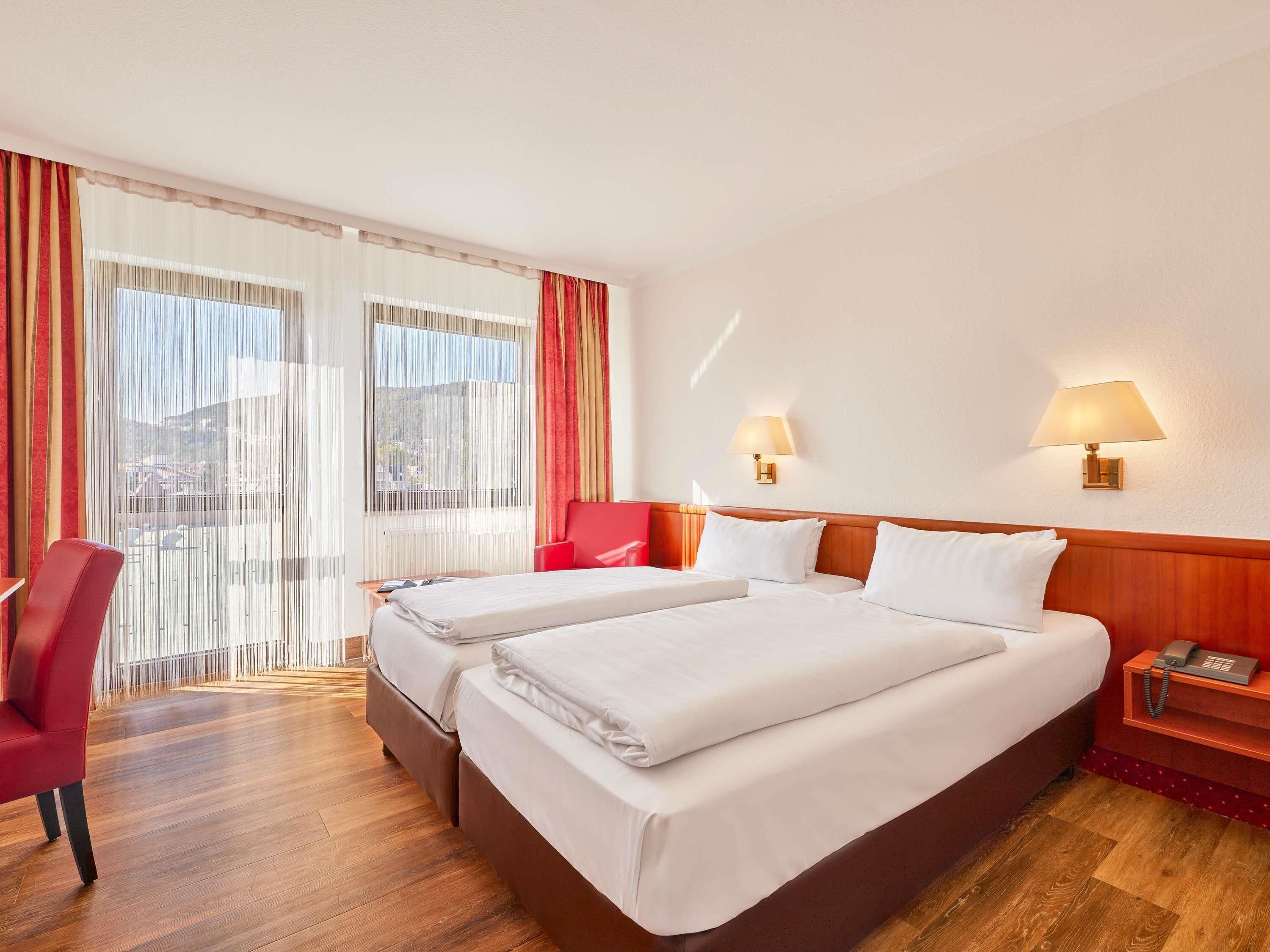 A hotel room with two beds, a red chair, a wooden nightstand with a phone, and a view through sheer curtains.