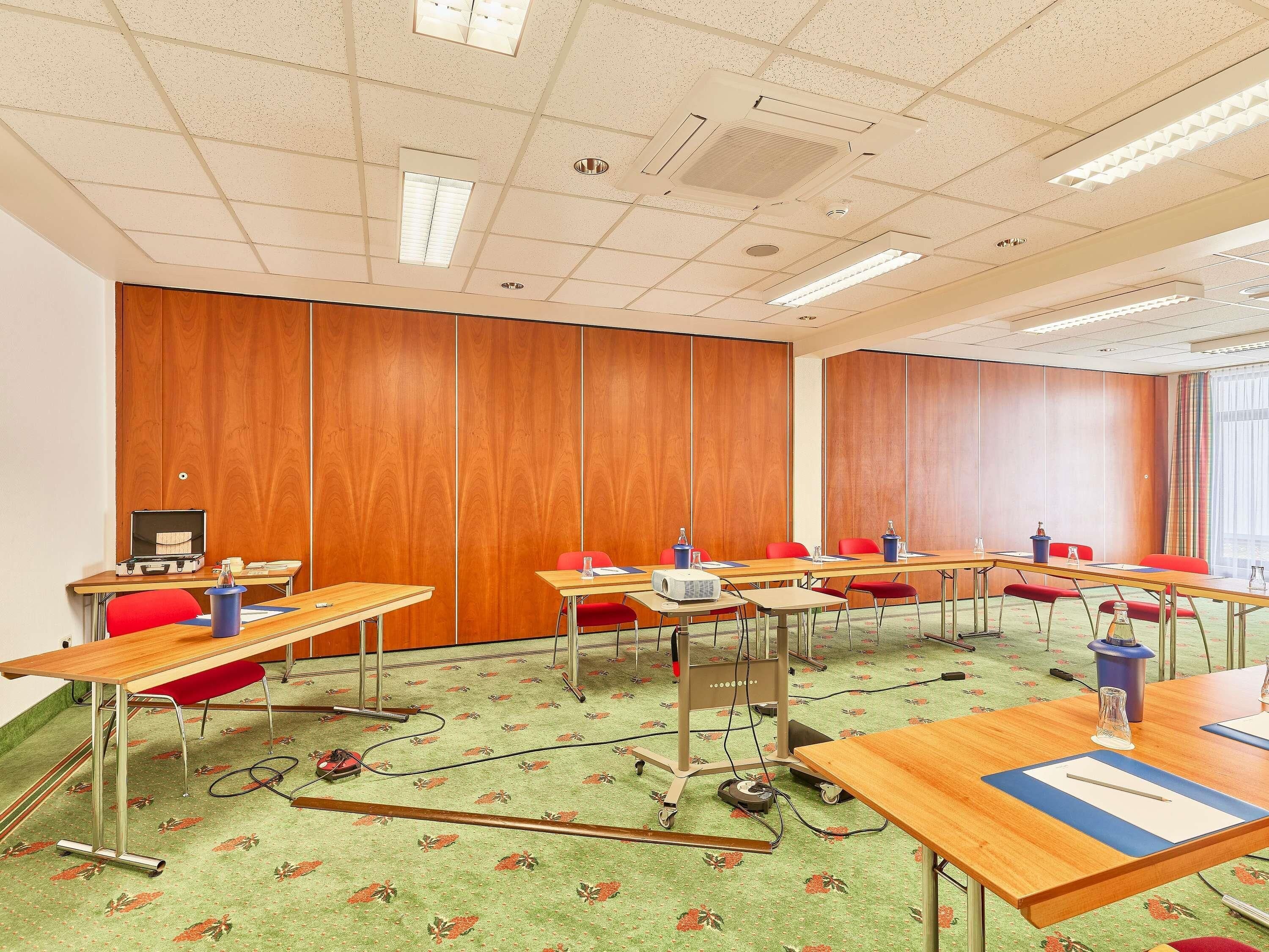 Un salón de conferencias con paredes de paneles de madera, varias mesas con sillas rojas y suelo alfombrado.