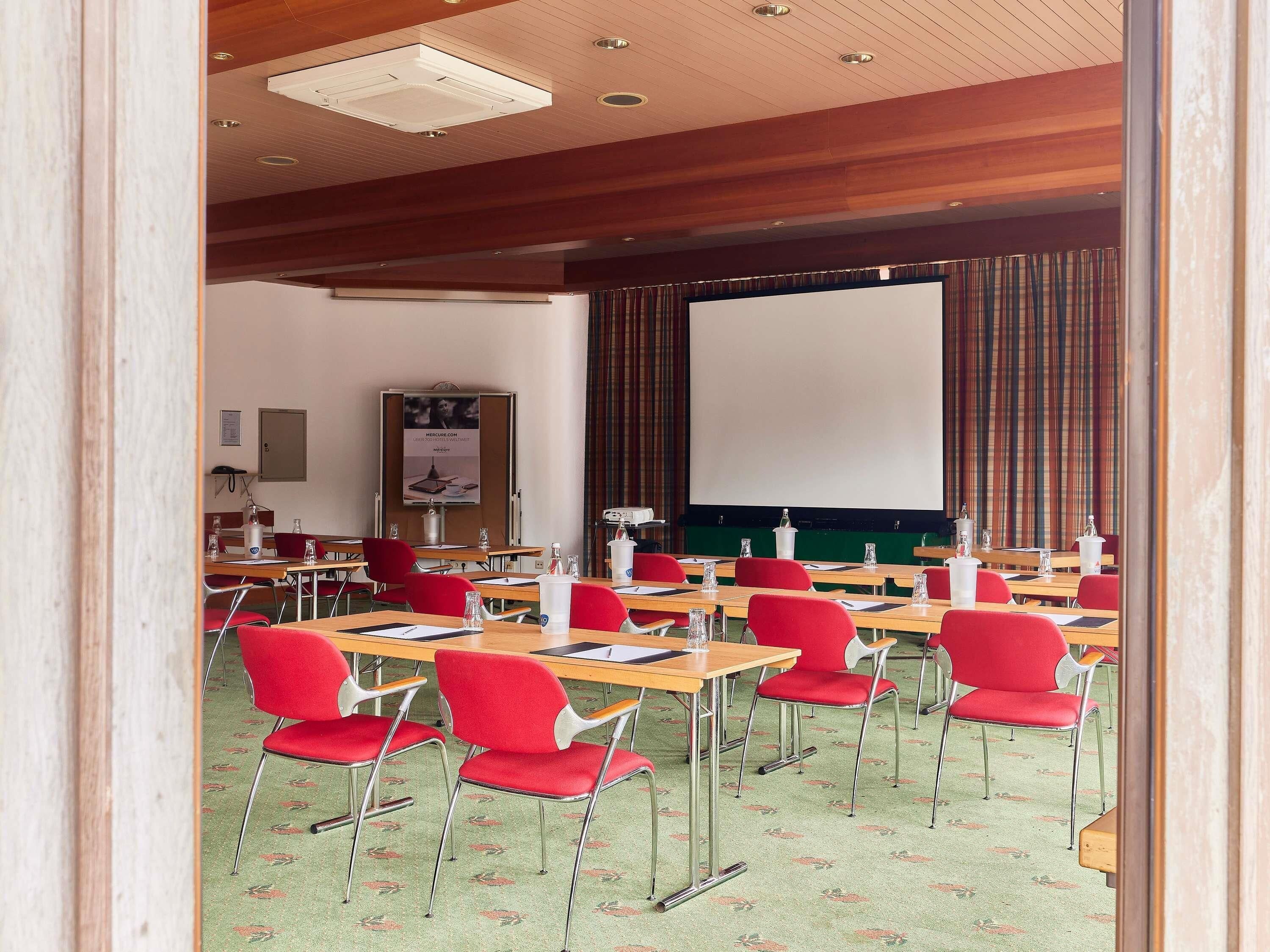 Un salón de conferencias con sillas rojas, una pantalla grande y un proyector.