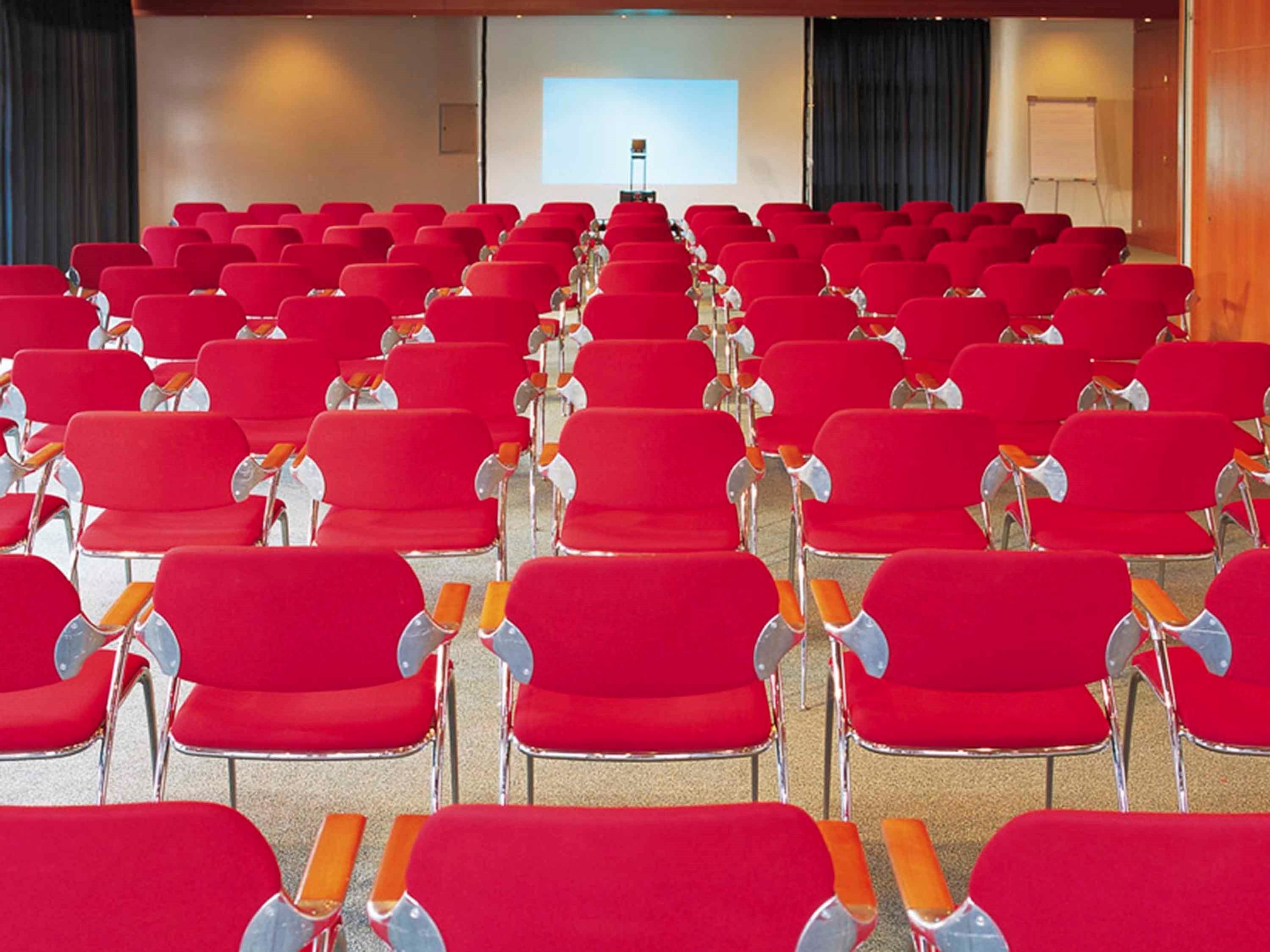 Une salle de conférence avec des rangées de chaises rouges et un grand écran à l’avant.