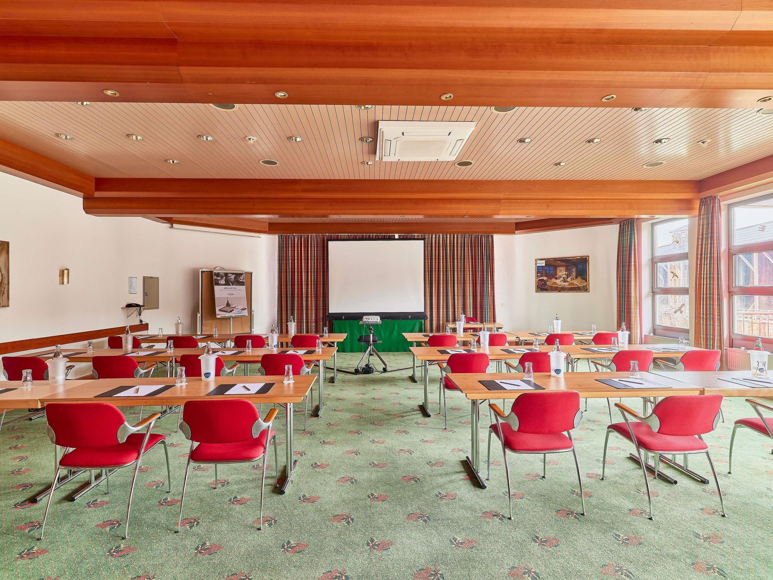 Sala de conferências com tetos de painel de madeira, cadeiras vermelhas e piso coberto de carpete.