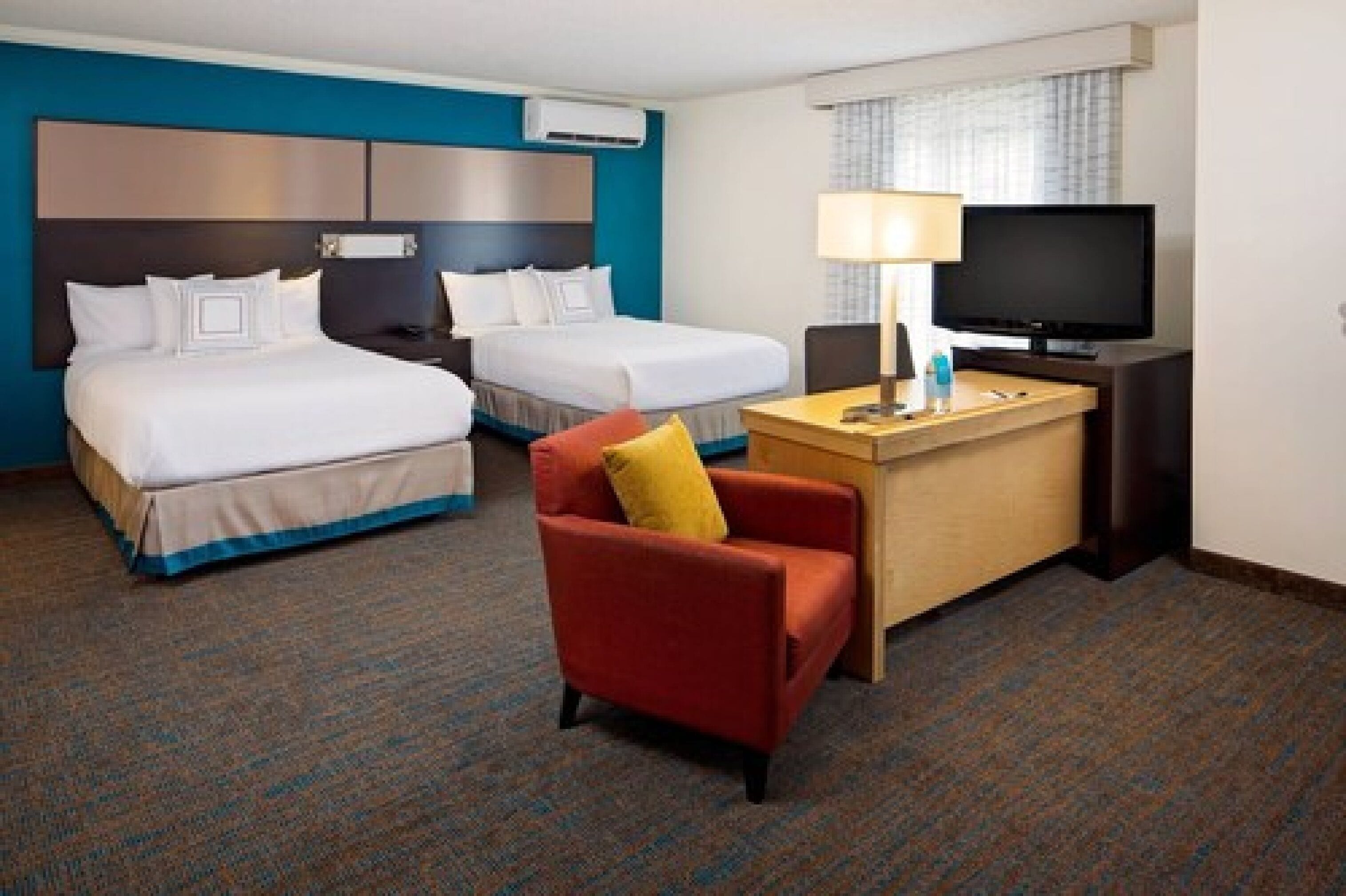 Ein Hotelzimmer mit zwei Betten, einem Schreibtisch mit Fernseher, einem Sessel und Teppichboden.