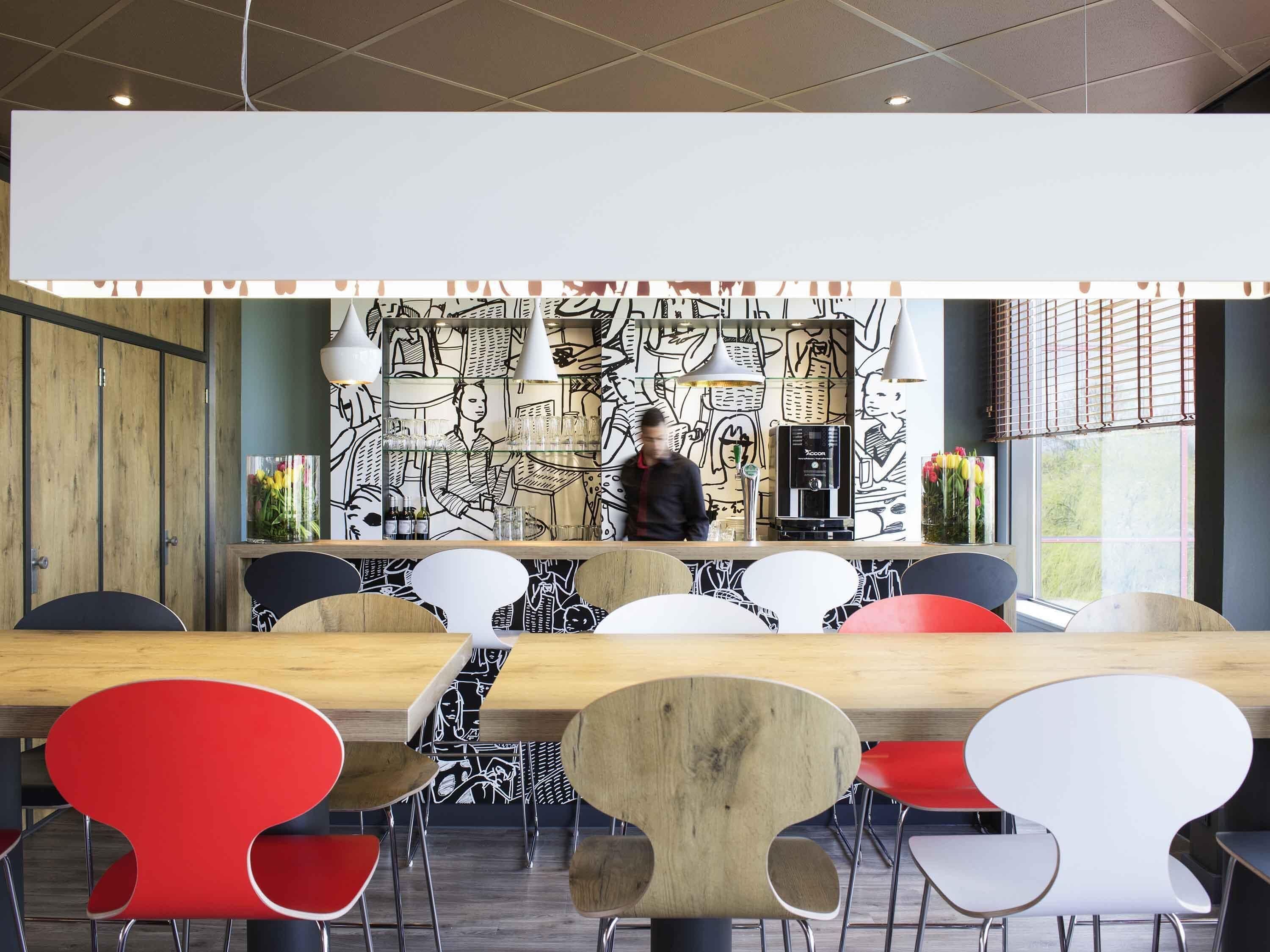 A modern bar area with a long wooden counter, black and white patterned wall art, and a mix of red and white chairs.