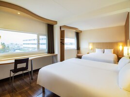 Habitación de hotel con dos camas, un escritorio, una silla y ventana con vistas a edificios.