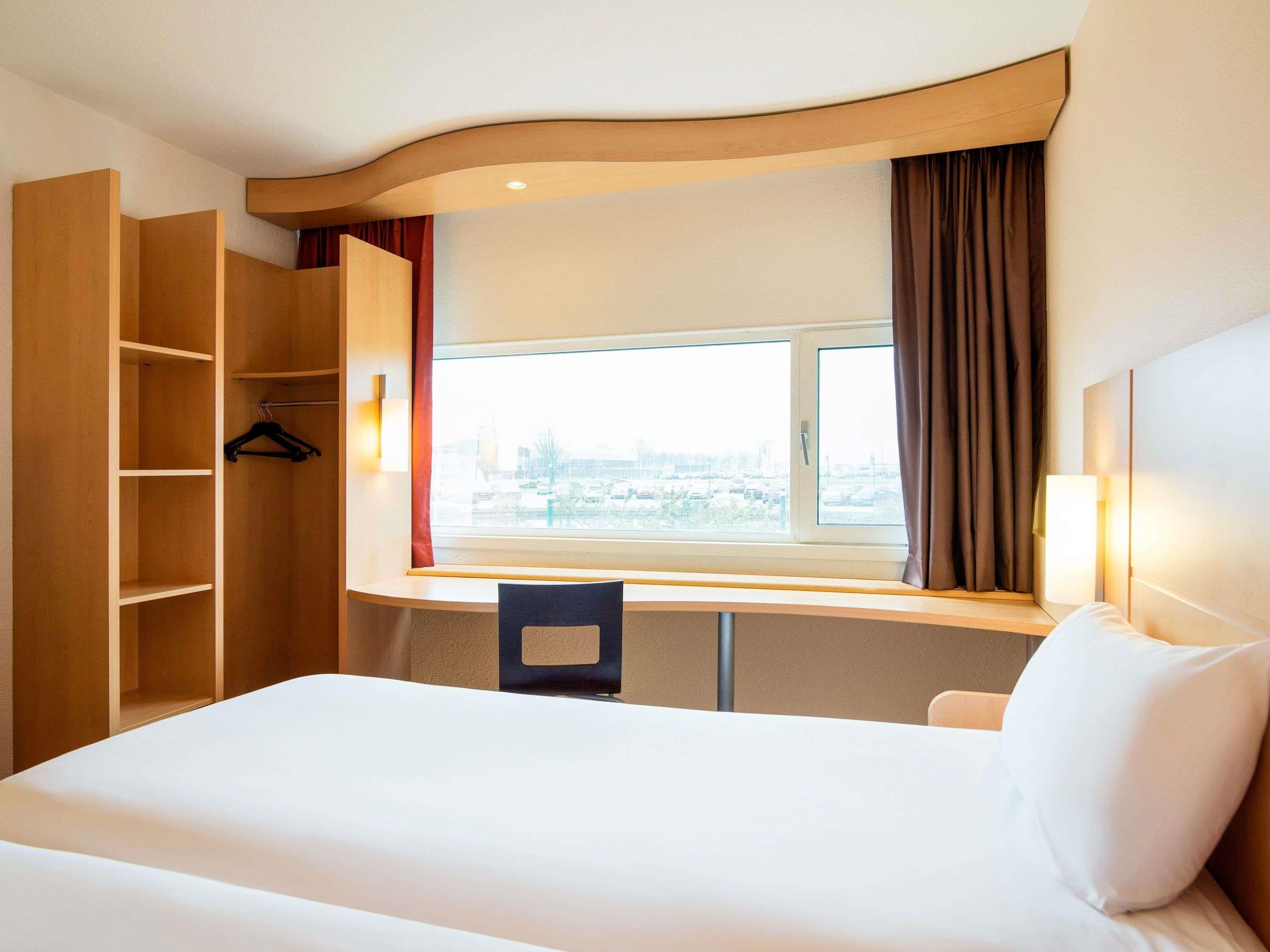 Una habitación de hotel con cama, escritorio, silla, armario y ventana con cortinas.