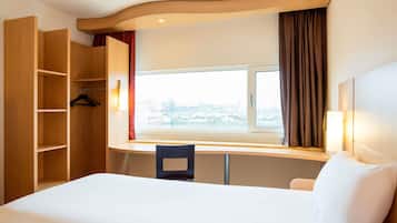 Una habitación de hotel con cama, escritorio, silla, armario y ventana con cortinas.