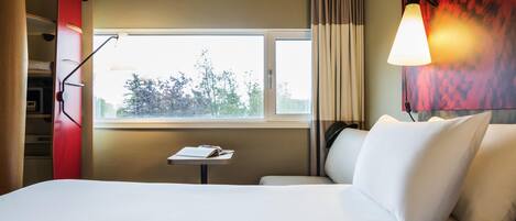 A hotel room with a bed, a small table with a book, a window with curtains, and a wall-mounted lamp.