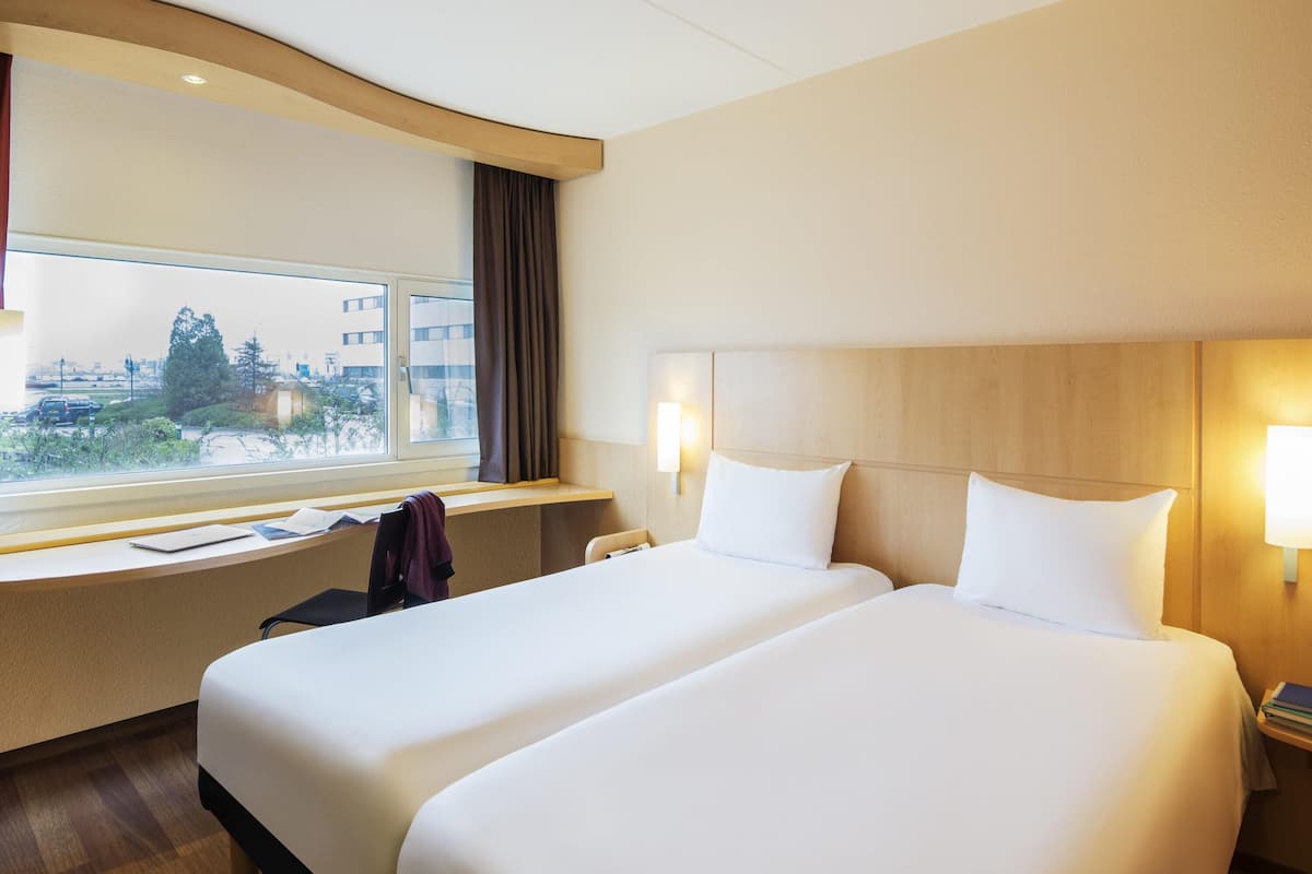 A hotel room with two beds, a desk, a window with a view of buildings and trees, and a lamp on the nightstand.