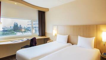 Habitación de hotel con dos camas, un escritorio, una ventana que ofrece vistas a edificios y árboles, y una lámpara en la mesita de noche.