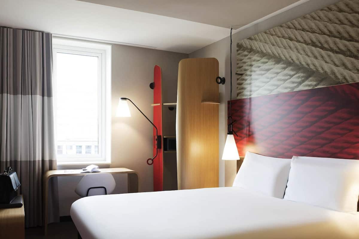 A hotel room with a large bed, a desk, a chair, a window with curtains, a red snowboard leaning against the wall, and a wall-mounted lamp.