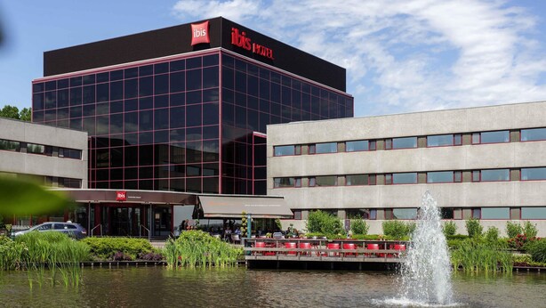 A modern hotel with a prominent glass facade, a water feature in front, and a clear sky. - Hotel ibis Schiphol Amsterdam Airport (Badhoevedorp)