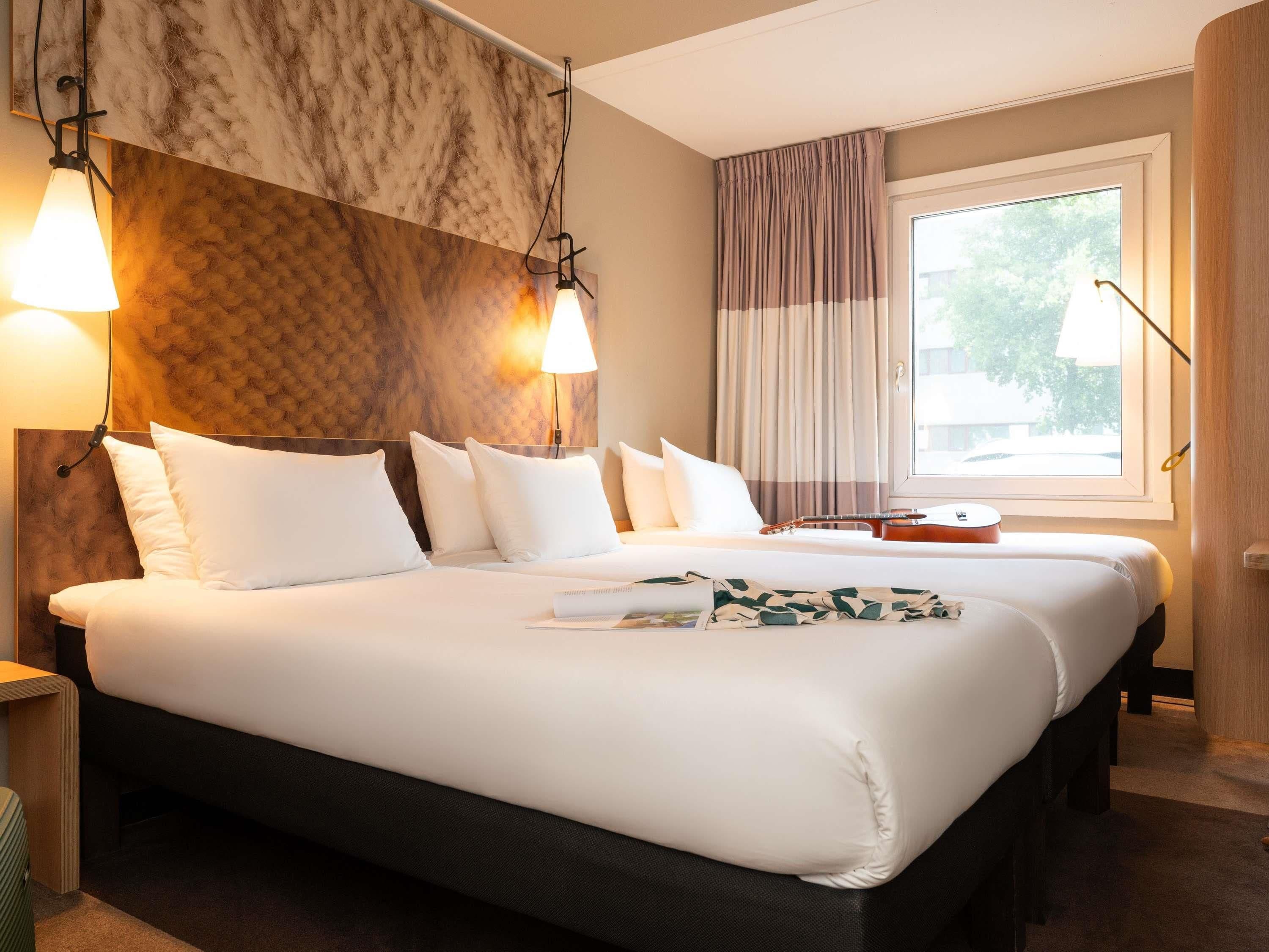 A hotel room with a large bed, two bedside lamps, a window with curtains, and a guitar on the bed.