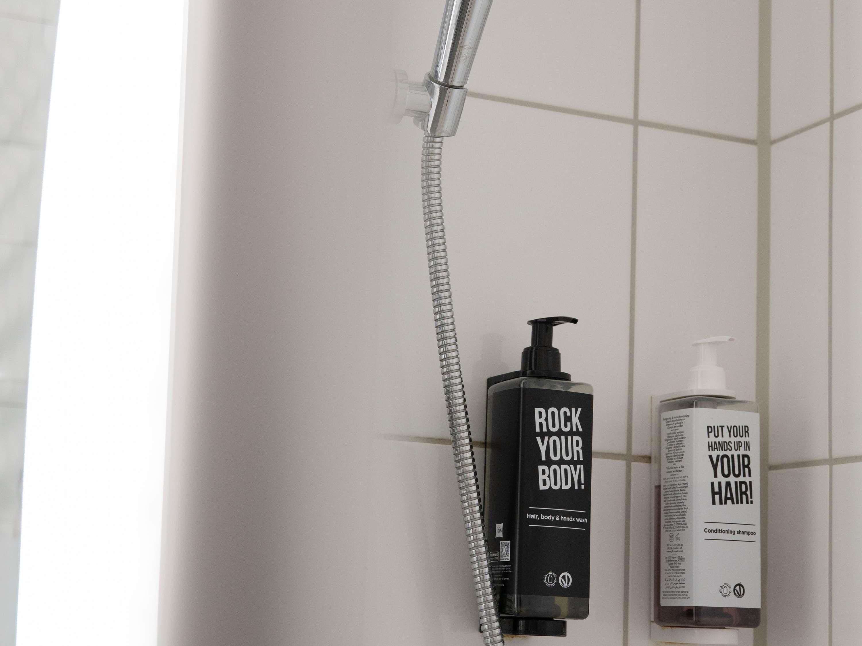 Un baño con una ducha, una botella negra etiquetada 'ROCK YOUR BODY!' y una botella blanca etiquetada 'PUT YOUR HANDS UP IN YOUR HAIR!'.