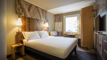 A hotel room with a large bed, two bedside lamps, a small table, a wardrobe, and a window with curtains.