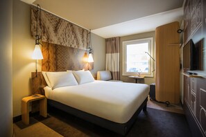 Habitación de hotel con una cama grande, dos lámparas de noche, una mesita, un armario y una ventana con cortinas.