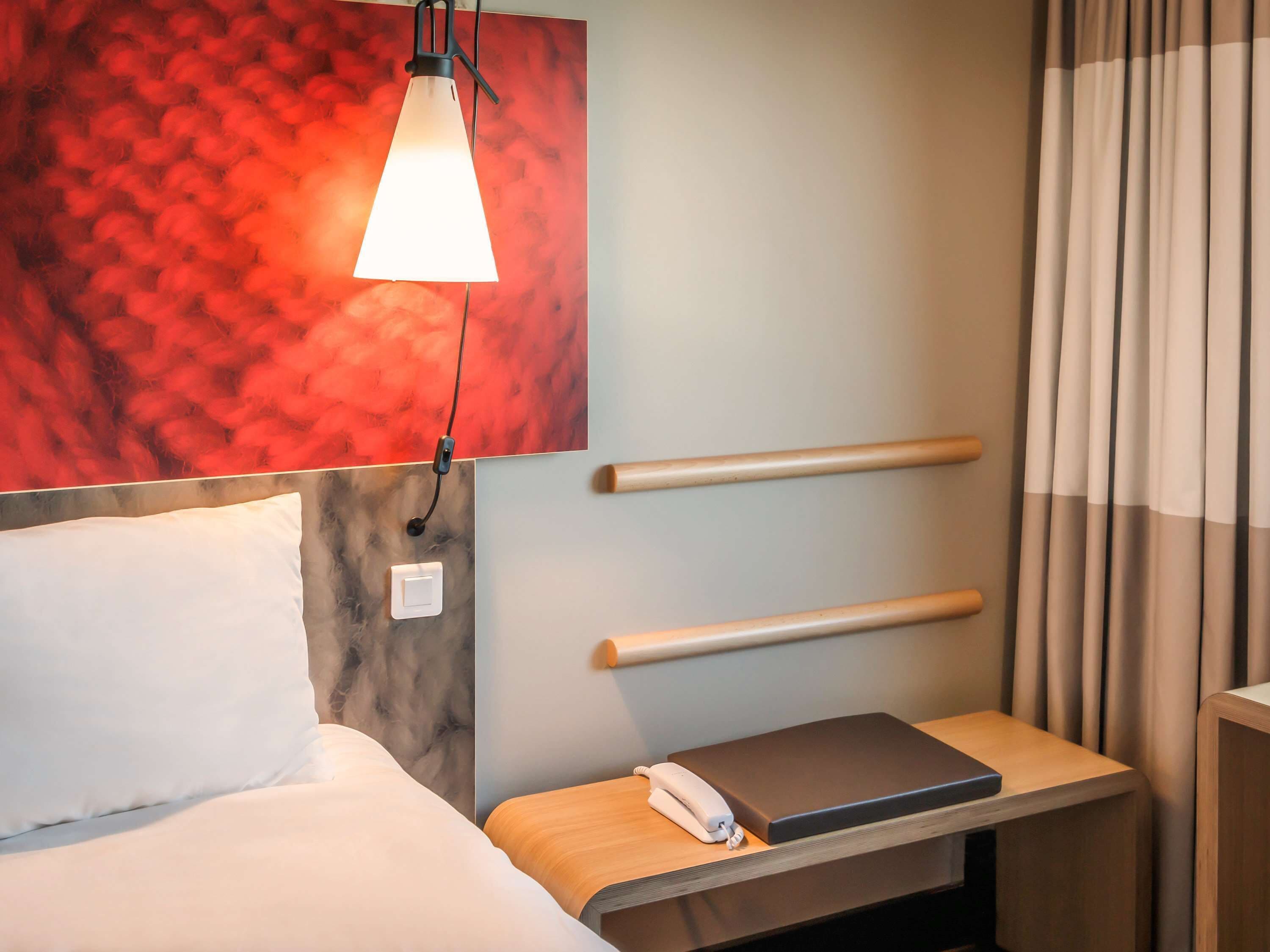 Habitación de hotel con una cama, una mesita de noche con un teléfono y un libro, una lámpara de pared y un cuadro abstracto rojo.