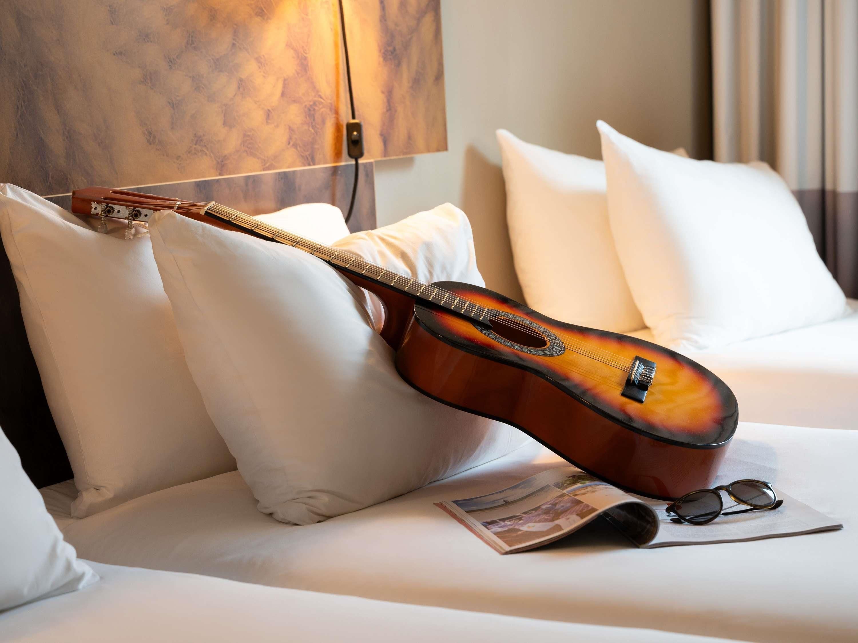 A guitar leaning against a white pillow on a bed with a magazine and glasses beside it.