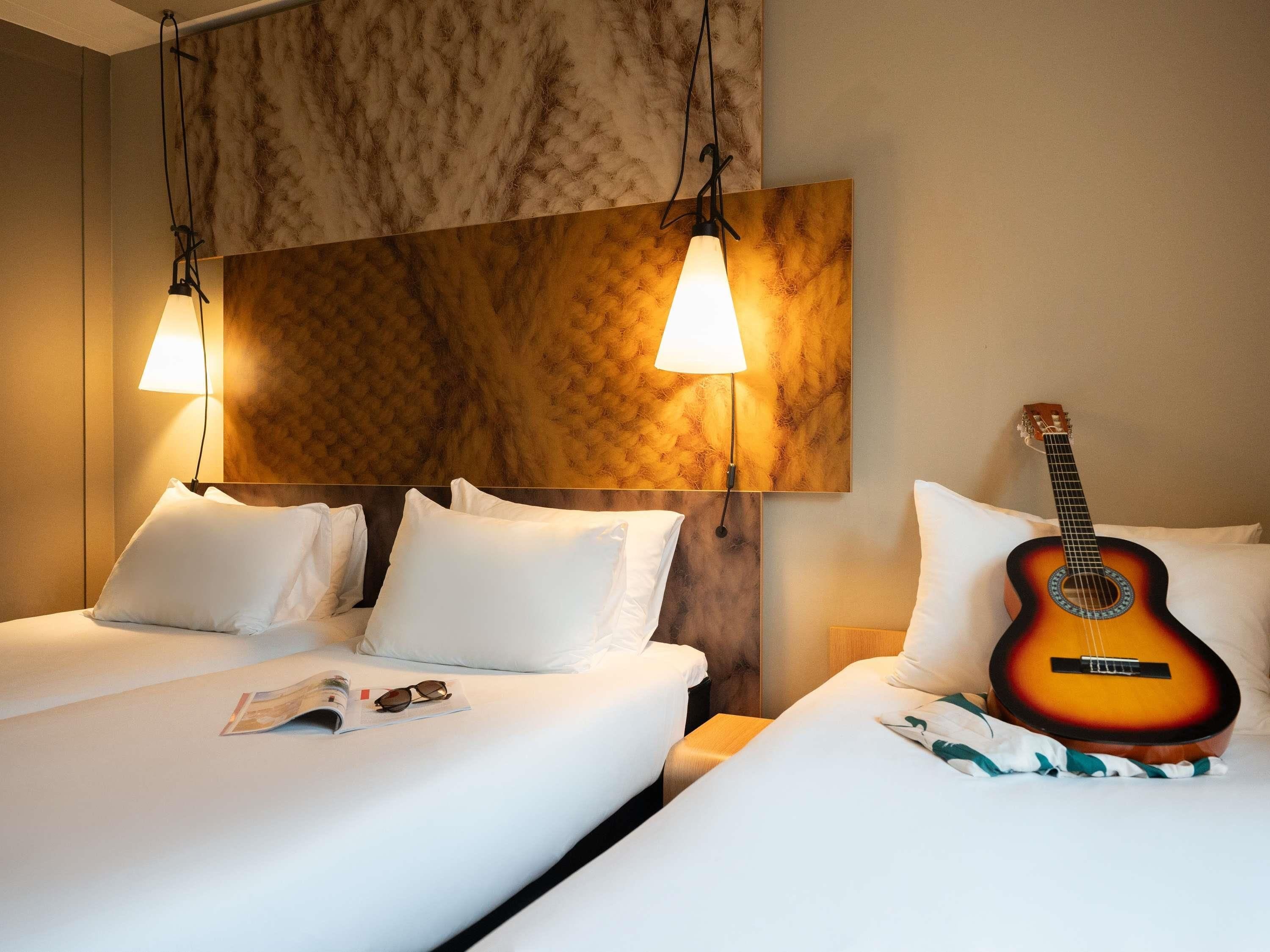 A hotel room with two beds, a guitar, and a wall-mounted lamp.