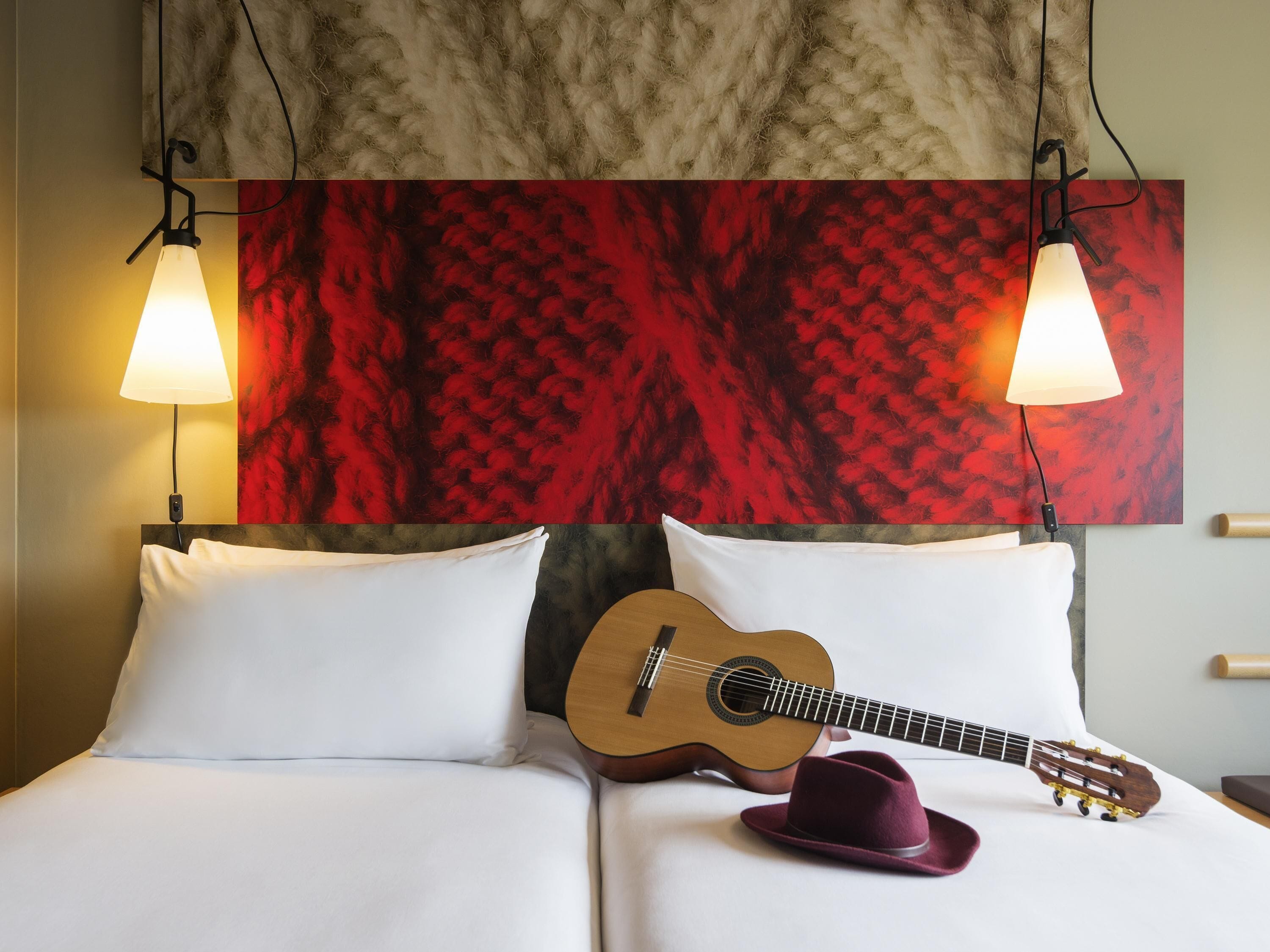 A hotel room with a bed, a guitar, and a hat on the bed. There are two wall-mounted lamps above the bed.