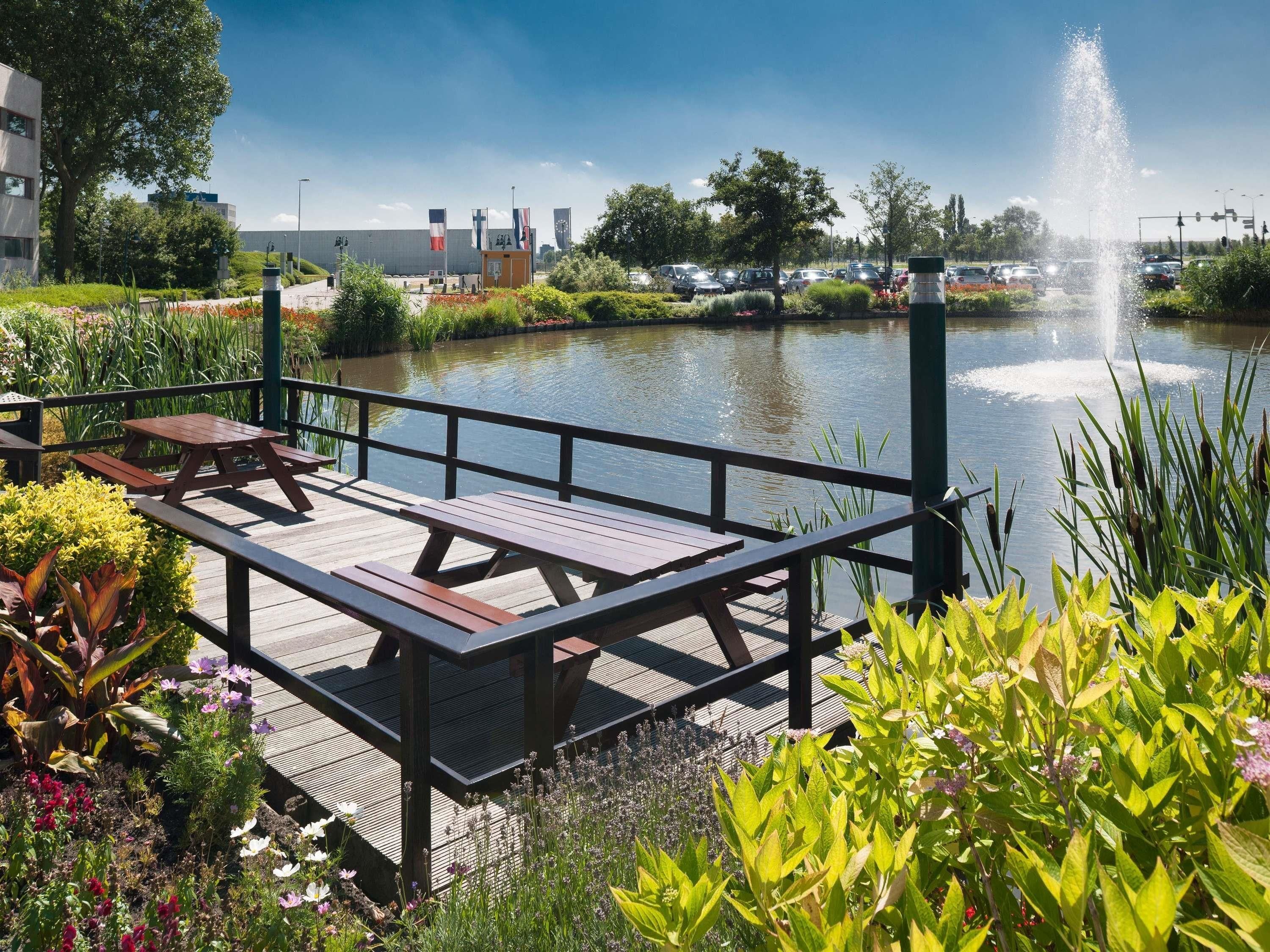 A wooden deck with picnic tables overlooking a calm water body, surrounded by lush greenery and a clear sky.