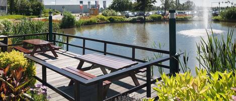 A wooden deck with picnic tables overlooking a calm water body, surrounded by lush greenery and a clear sky.