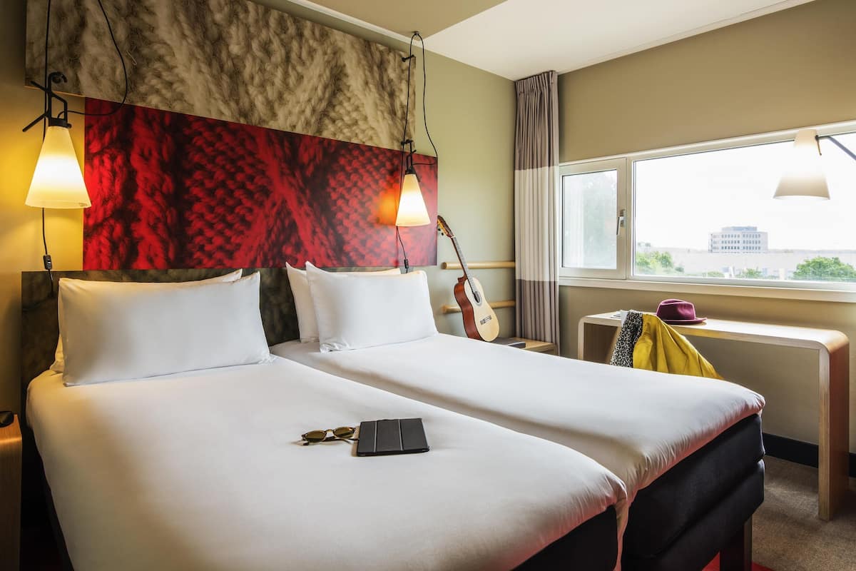 A hotel room with a bed, two bedside lamps, a guitar, a window with a view, and a small table with a hat and jacket.