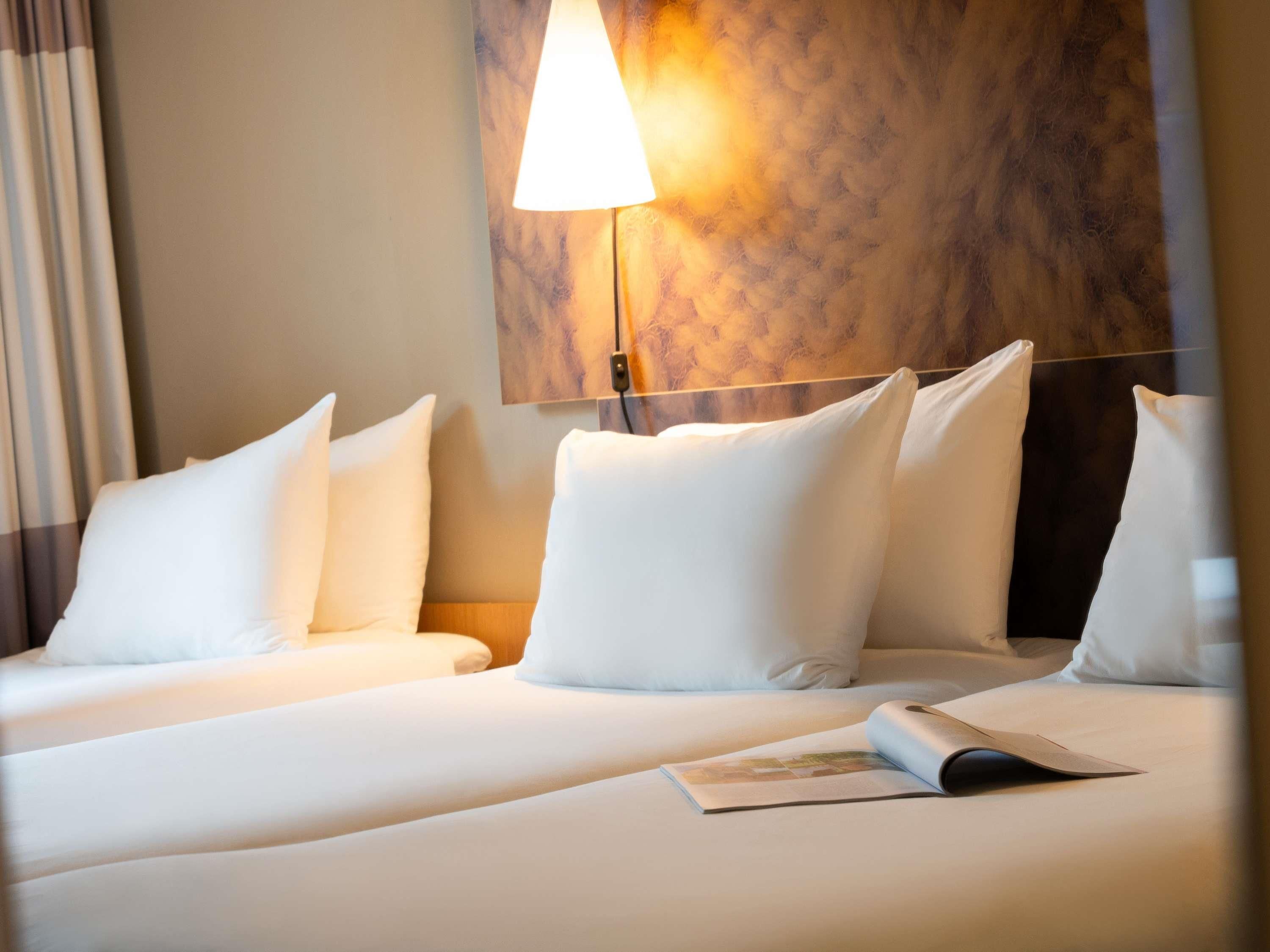 A hotel room with two beds, a wall-mounted lamp, and a magazine on the bed.