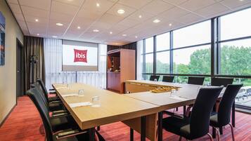 Un salón de conferencias con una larga mesa, sillas, una pantalla de proyector mostrando "ibis Hotels" y grandes ventanales con vistas a zonas verdes.