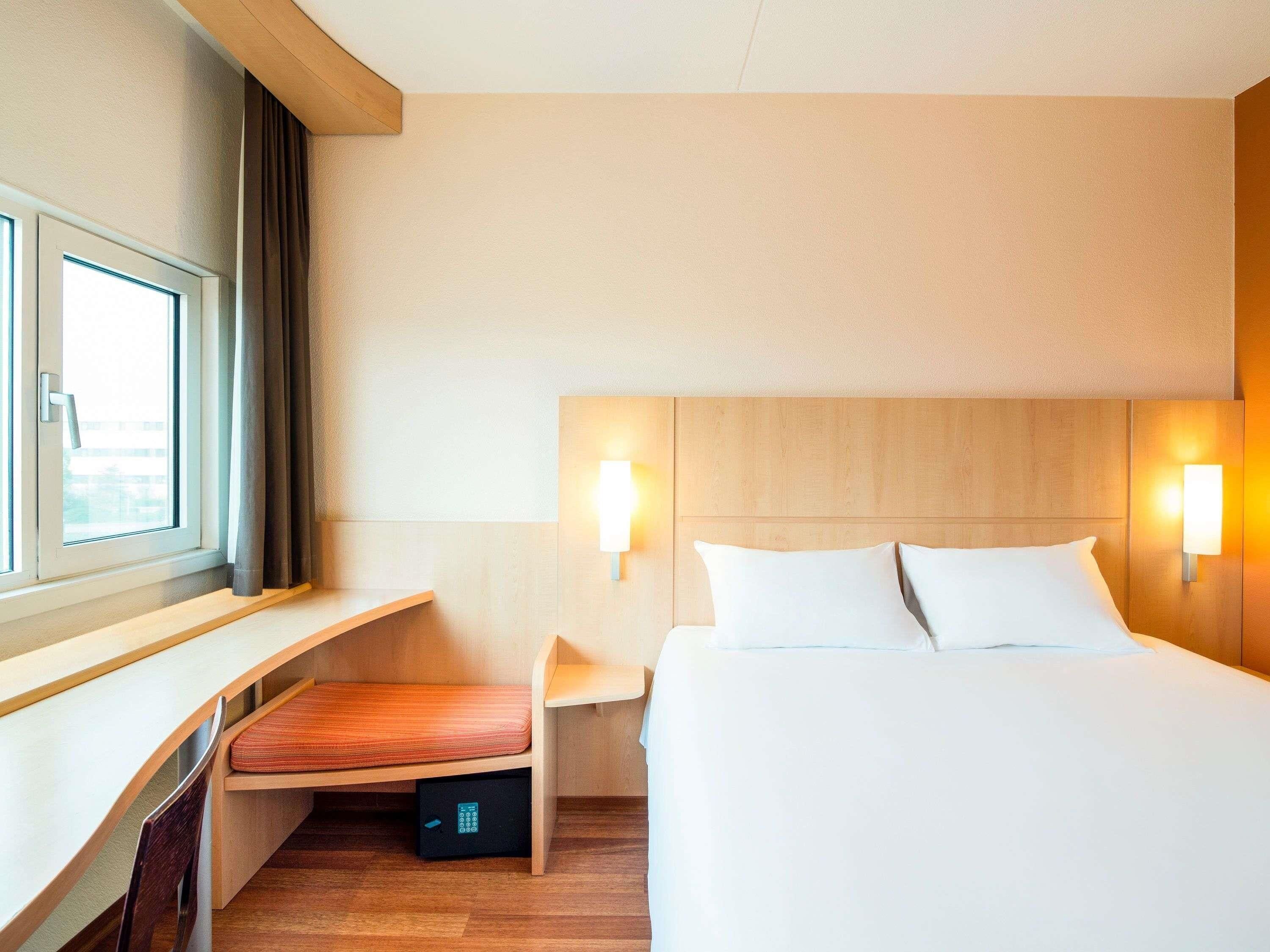 Ett hotellrum med en säng, sängbord, ett fönster med gardiner, ett skrivbord med en stol och ett litet förvaringsskåp.