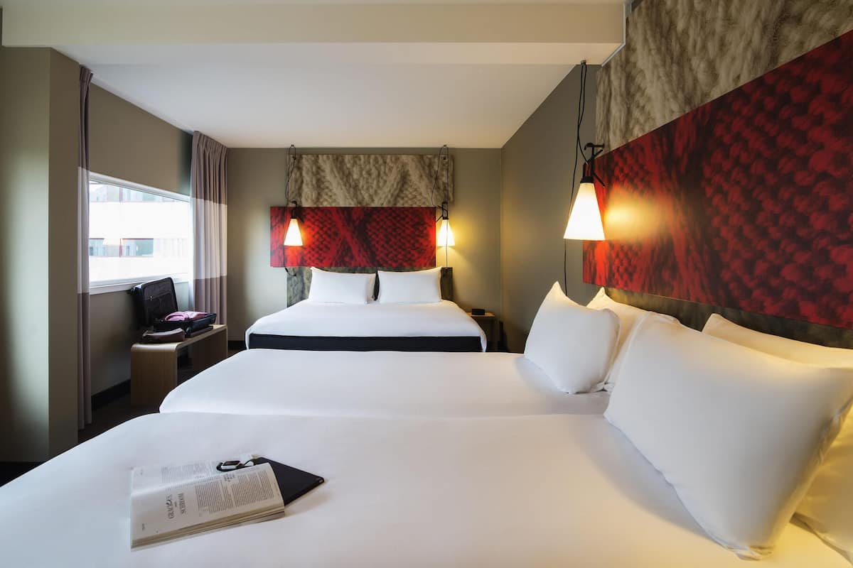 A hotel room with two beds, a nightstand, a lamp, and a window with curtains.
