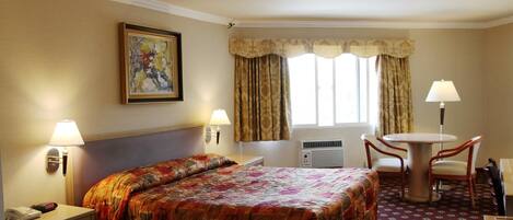 A hotel room with a large bed, a desk with a lamp, a chair, and a table. There is a painting on the wall, a window with curtains, and an air conditioning unit.