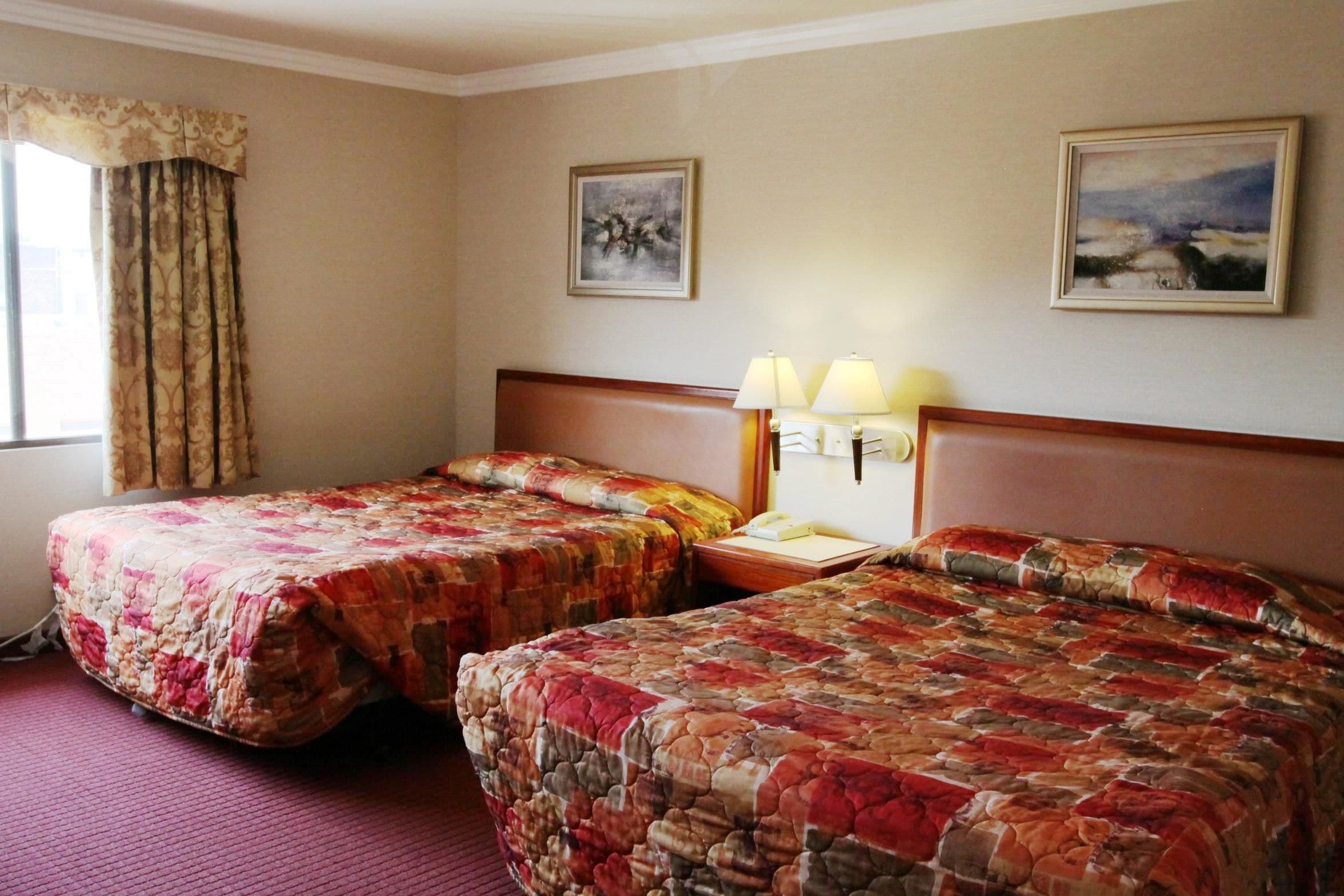 A hotel room with two beds, a headboard, a nightstand with lamps, and two wall-mounted paintings.