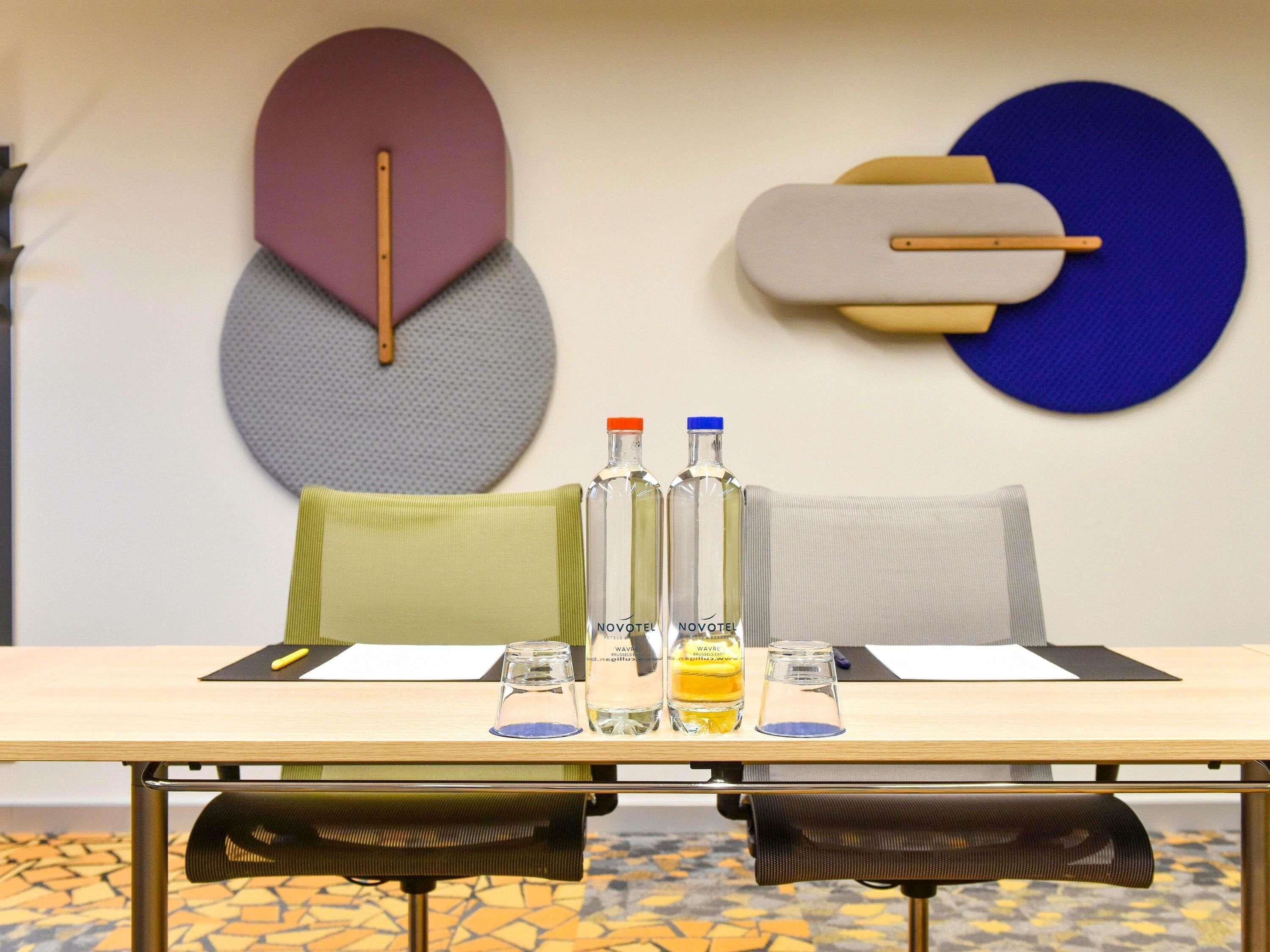 A hotel room with a wooden table, two bottles of Novotel Wavre, a pair of glasses, and a patterned floor.