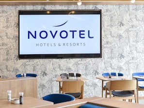 Ein Innenraum eines Novotel Hotels mit einem großen Bildschirm, der das Novotel-Logo zeigt, Holzmöbeln und einer Wand mit tropischem Blattmuster.