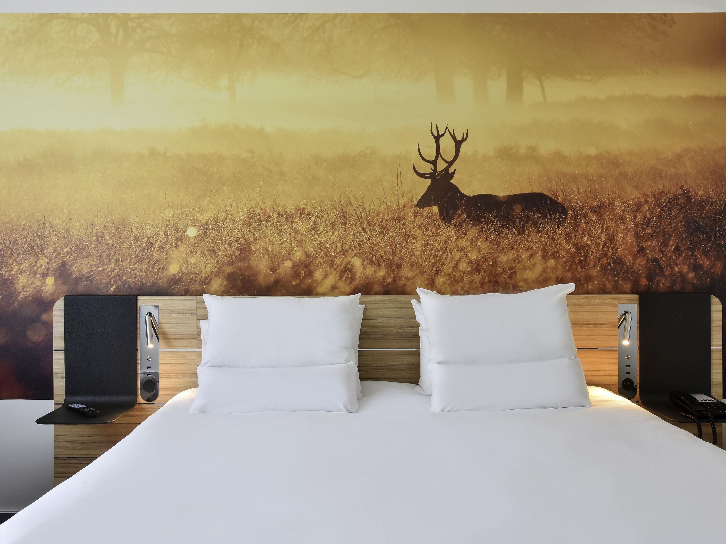Een hotelkamer met een groot bed, twee witte kussens, een hoofdbord met een vastgezet telefoon en een muurschildering van een herten in een veld.